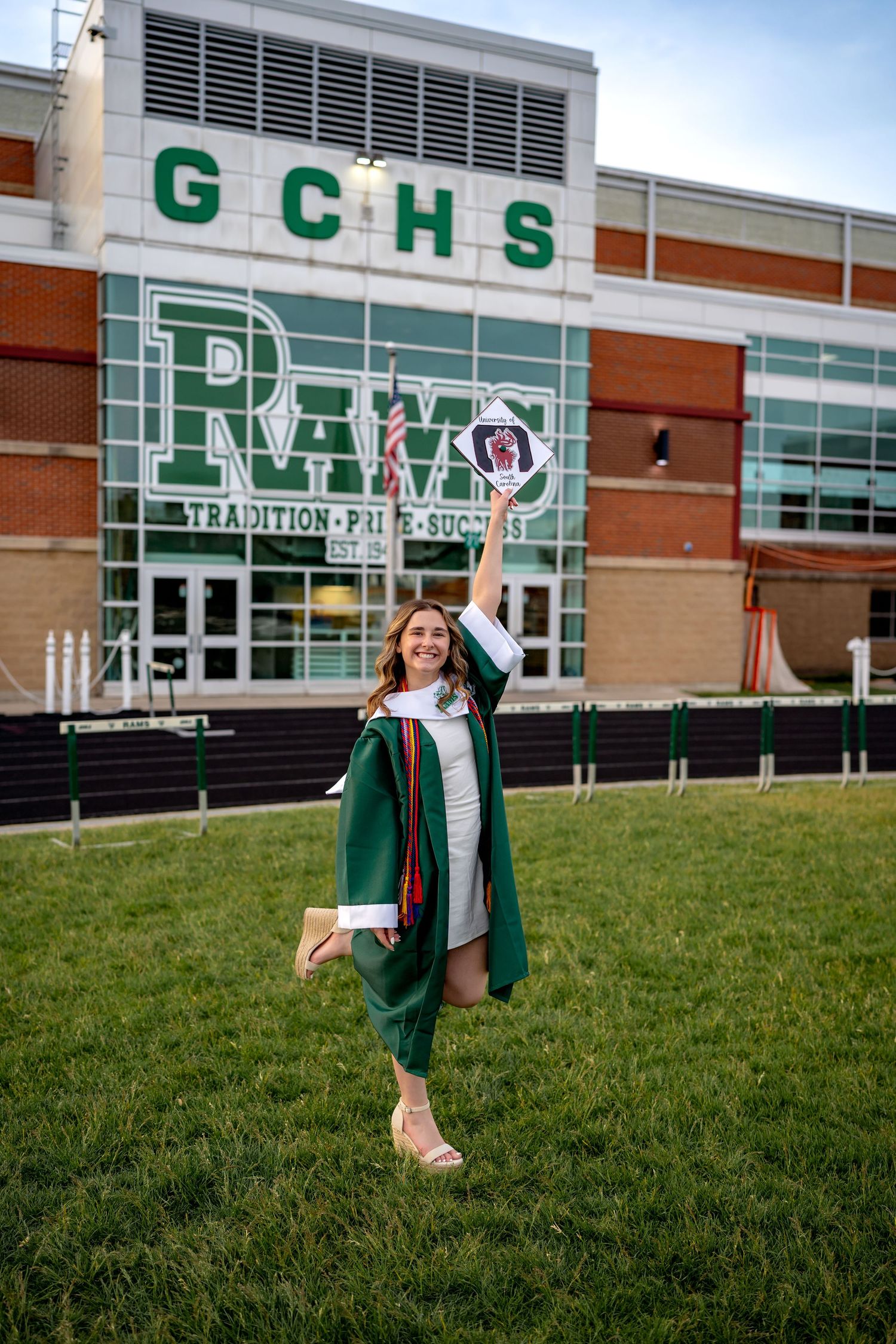 Graduate in green cap and gown celebrates on lawn in front of high school building with GCHS signage.