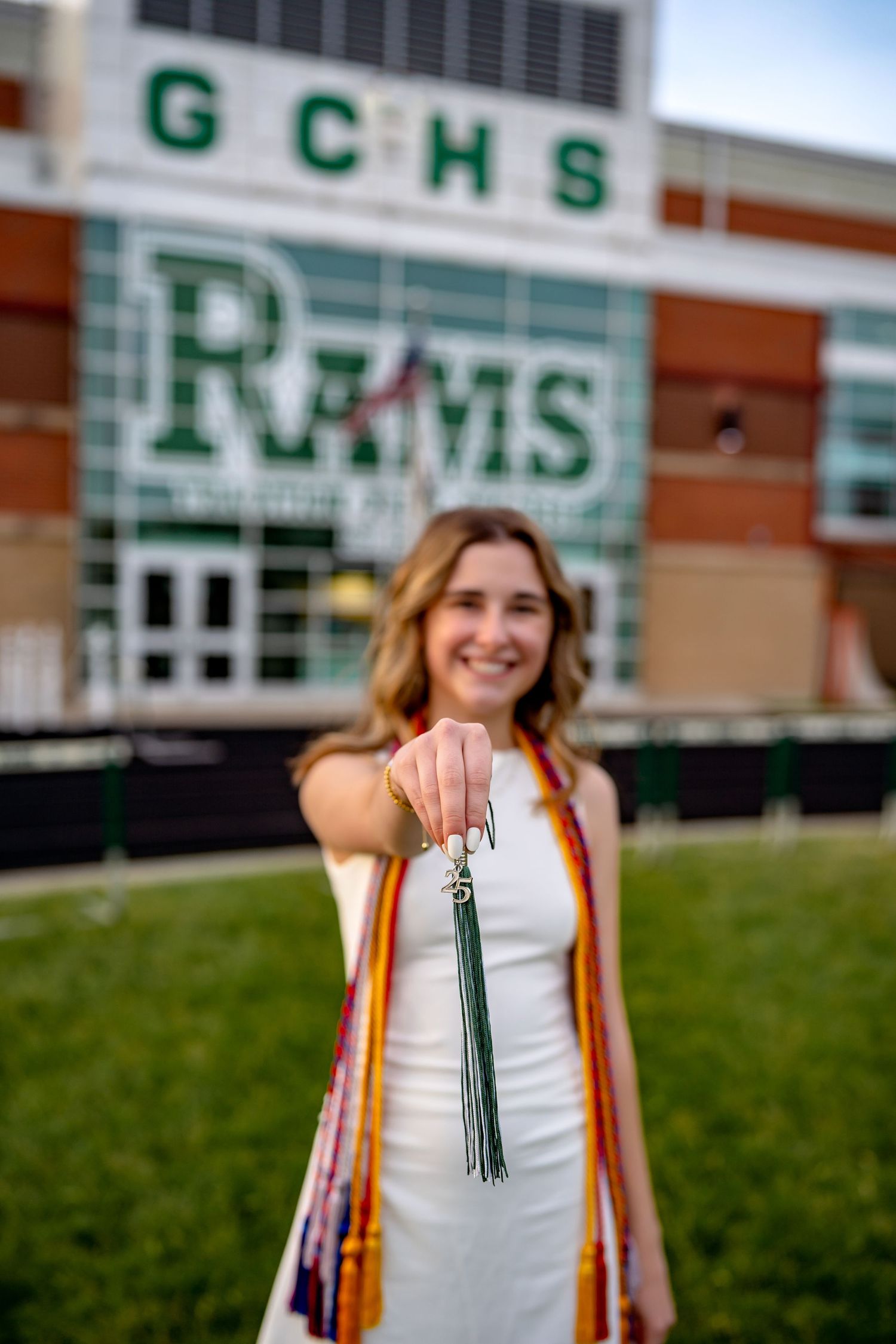 Graduate poses in white dress with honor cords in front of GCHS high school entrance.