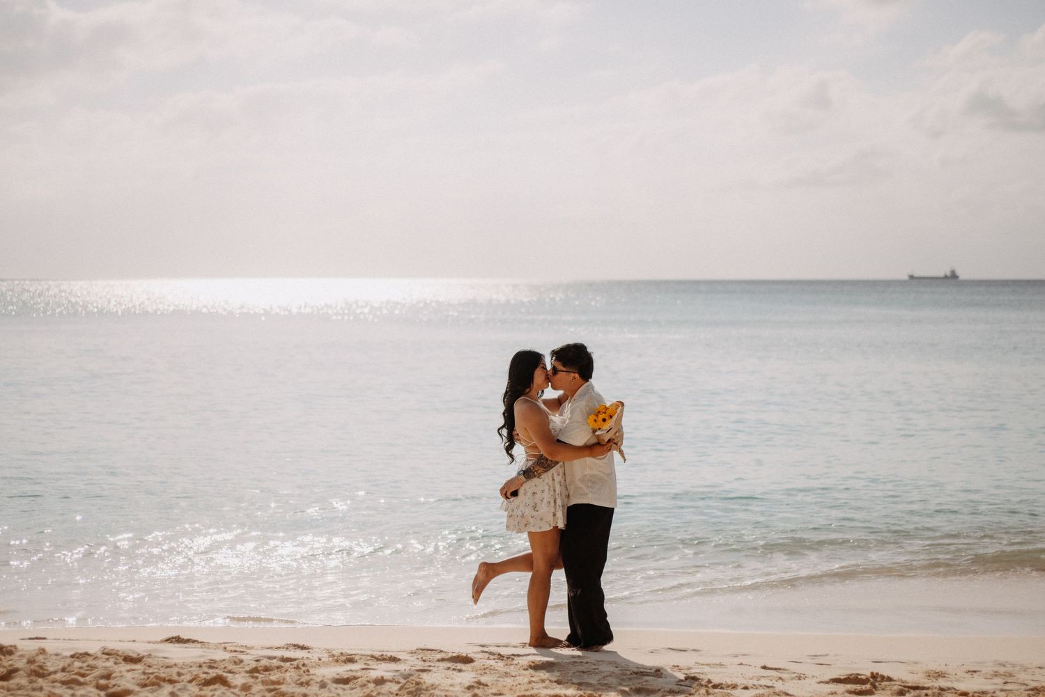 A romantic couple shares an intimate moment on a serene beach with gentle waves and cloudy skies in the background.