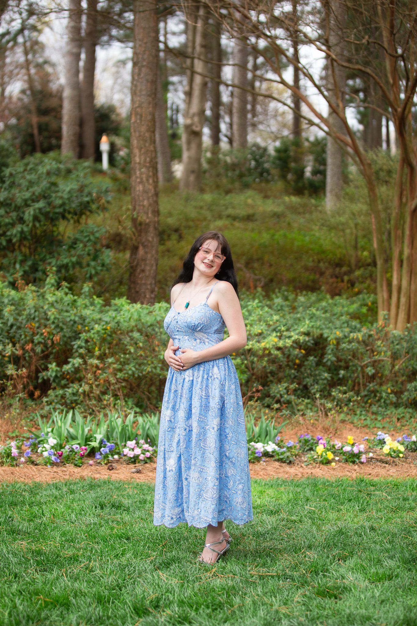 Senior Grad in blue floral sundress standing on green lawn surrounded by trees and colorful flowers.