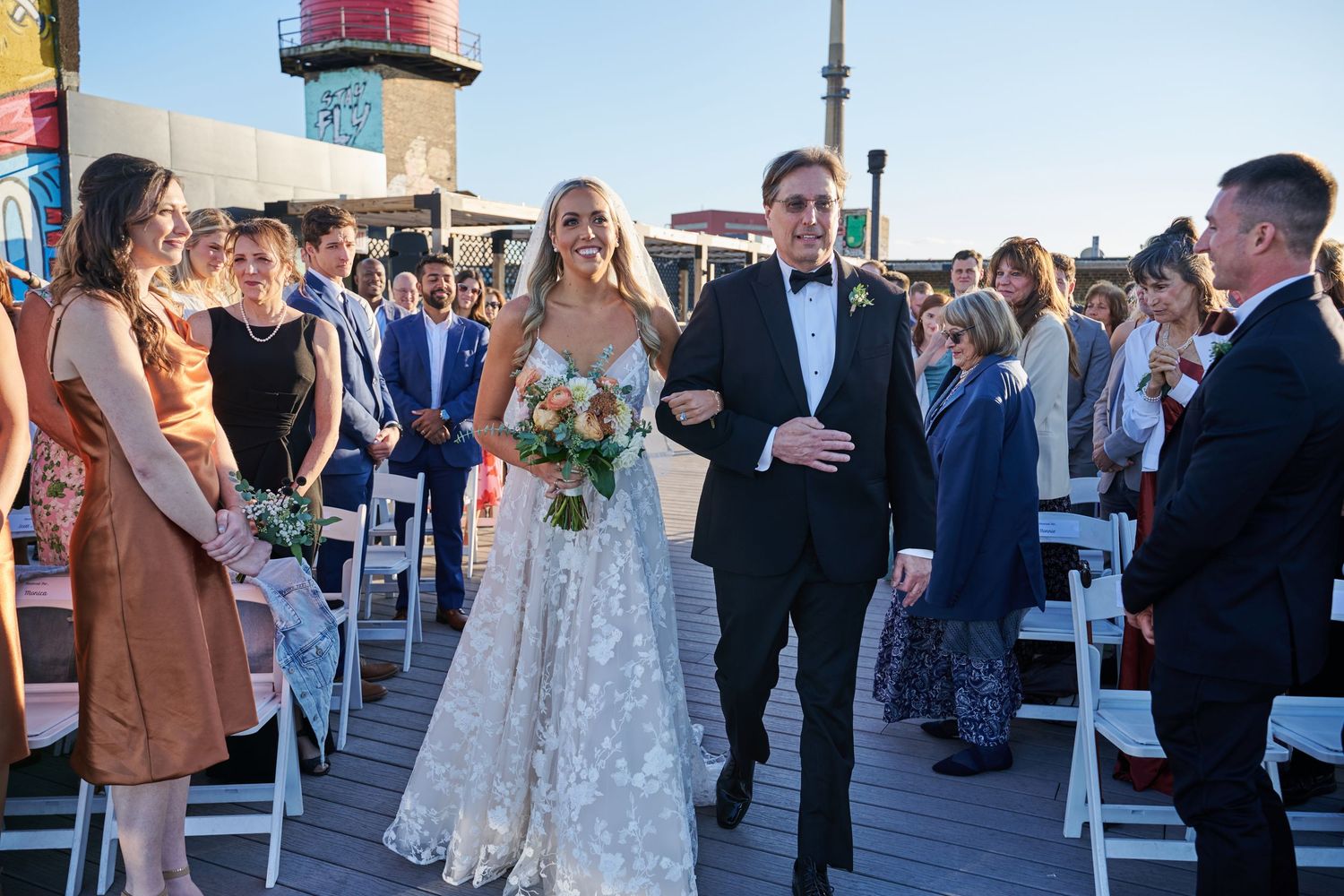Wedding guests gather on a sunny outdoor deck with string lights and a lighthouse in the background during a celebration.