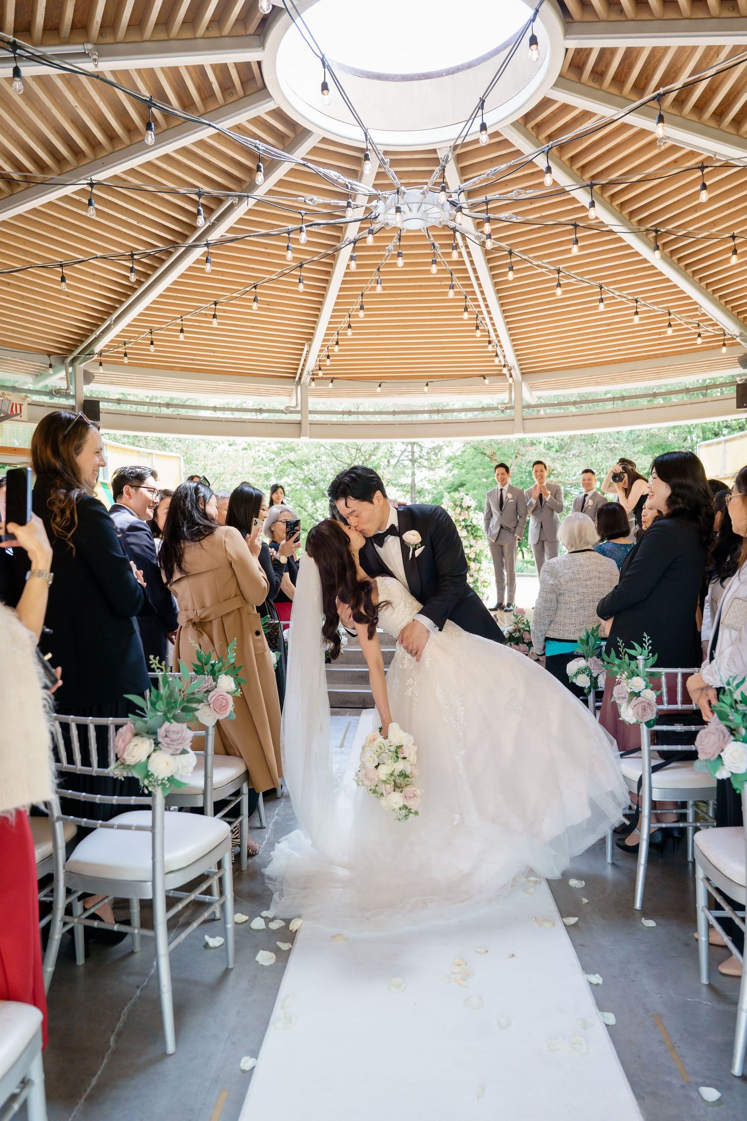 A couple shares a romantic dip during their wedding ceremony in an elegant outdoor gazebo with wooden beams and white decor.Queen Elizabeth
