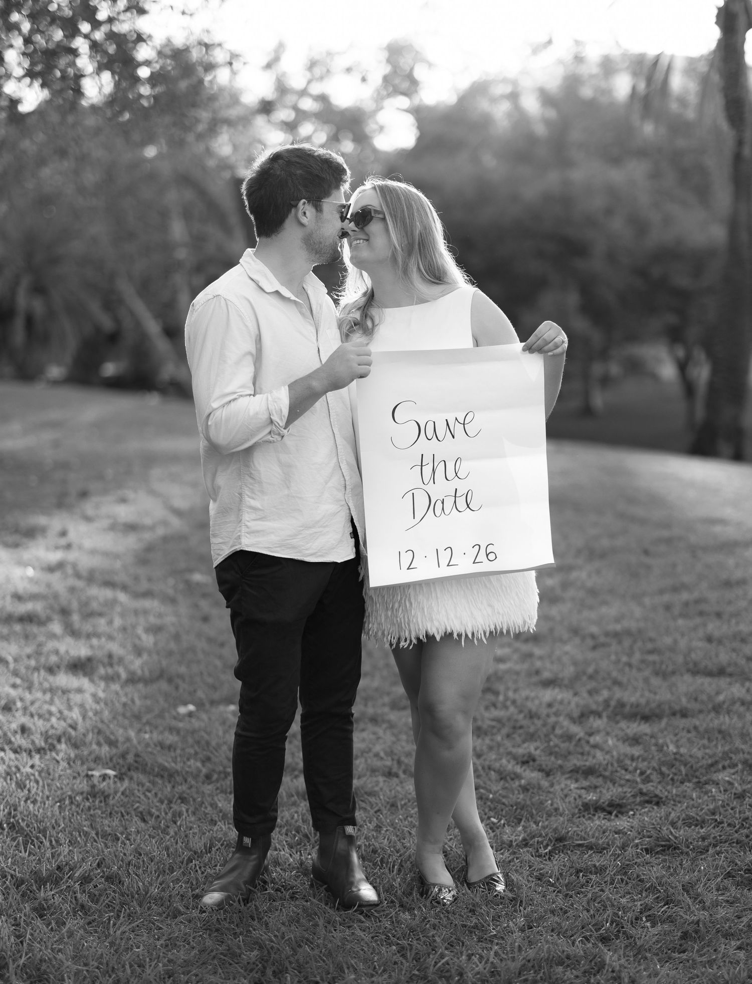 A couple in white outfits holds up handwritten Save the Date cards during a golden sunset photoshoot outdoors.