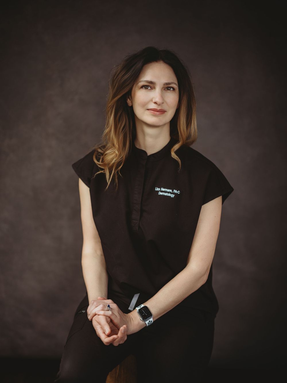 Professional portrait of someone wearing a black scrub top with short sleeves posing with clasped hands against dark background.