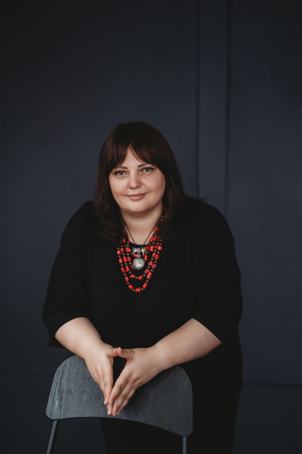 Professional portrait of someone wearing a black top with layered red beaded necklaces against a dark background.