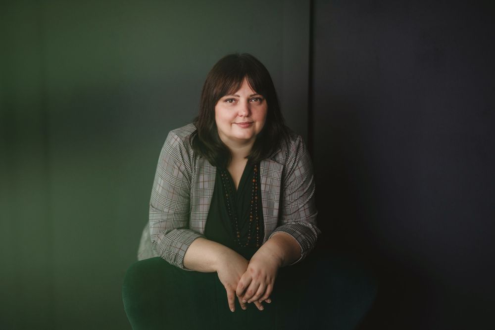 Professional indoor portrait of a person sitting against a dark green background in business casual attire.