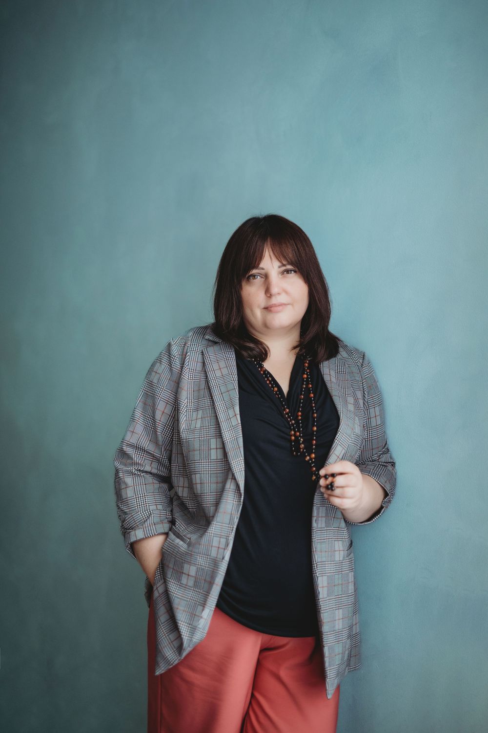 Business casual portrait of someone in a gray blazer and coral pants against a teal wall.