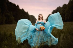 A flowing turquoise maternity gown billows dramatically in a meadow at sunset as sunflowers are held against the dress.