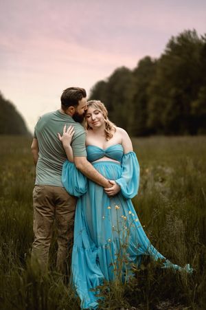A couple embraces tenderly in a grassy meadow, with the person in a flowing turquoise dress glowing in the soft evening light.