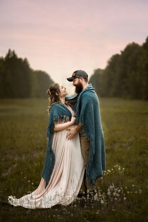 A romantic maternity photo in a meadow at dusk shows a couple embracing while wearing matching teal wraps.