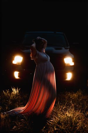 A silhouetted figure in a flowing dress stands against the glow of car headlights in the darkness.