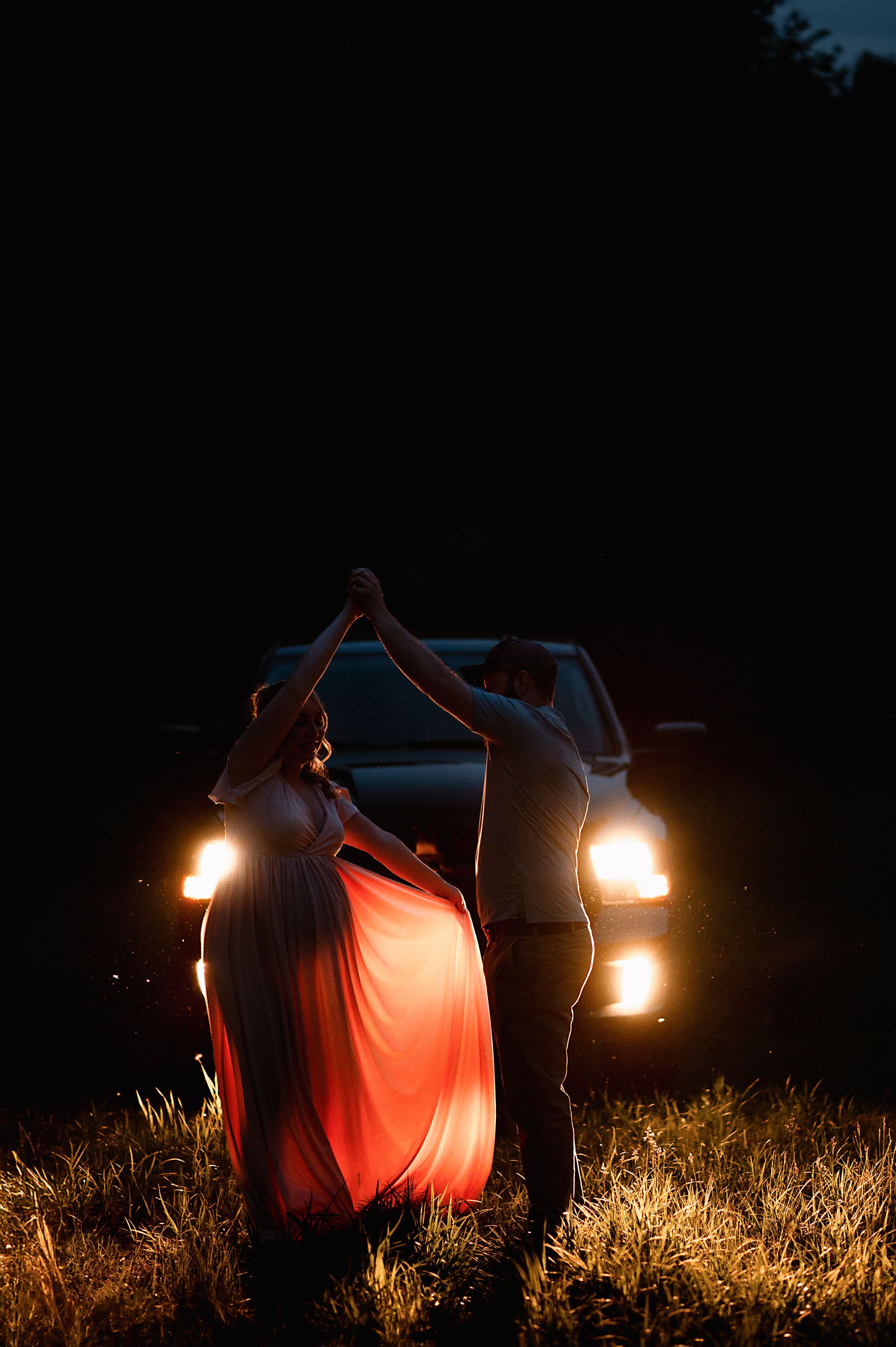 Romantic silhouette of two dancers illuminated by car headlights in a dark field at night.