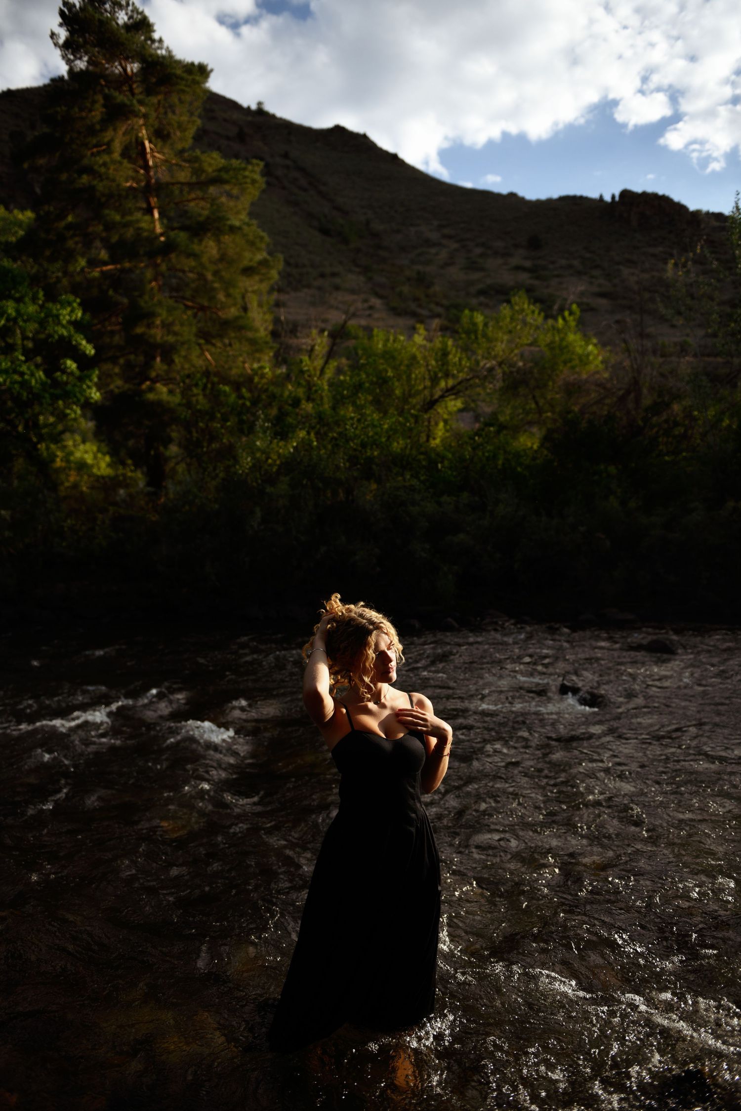 Silhouette of someone in a black dress standing in a rushing river with dramatic mountain cliffs and trees in background.