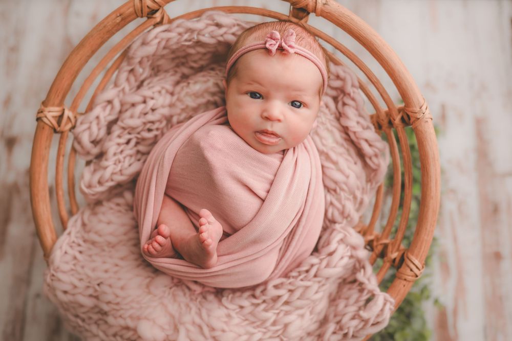 Baby wrapped in soft pink swaddle lying in circular wooden prop basket with textured blanket and greenery.