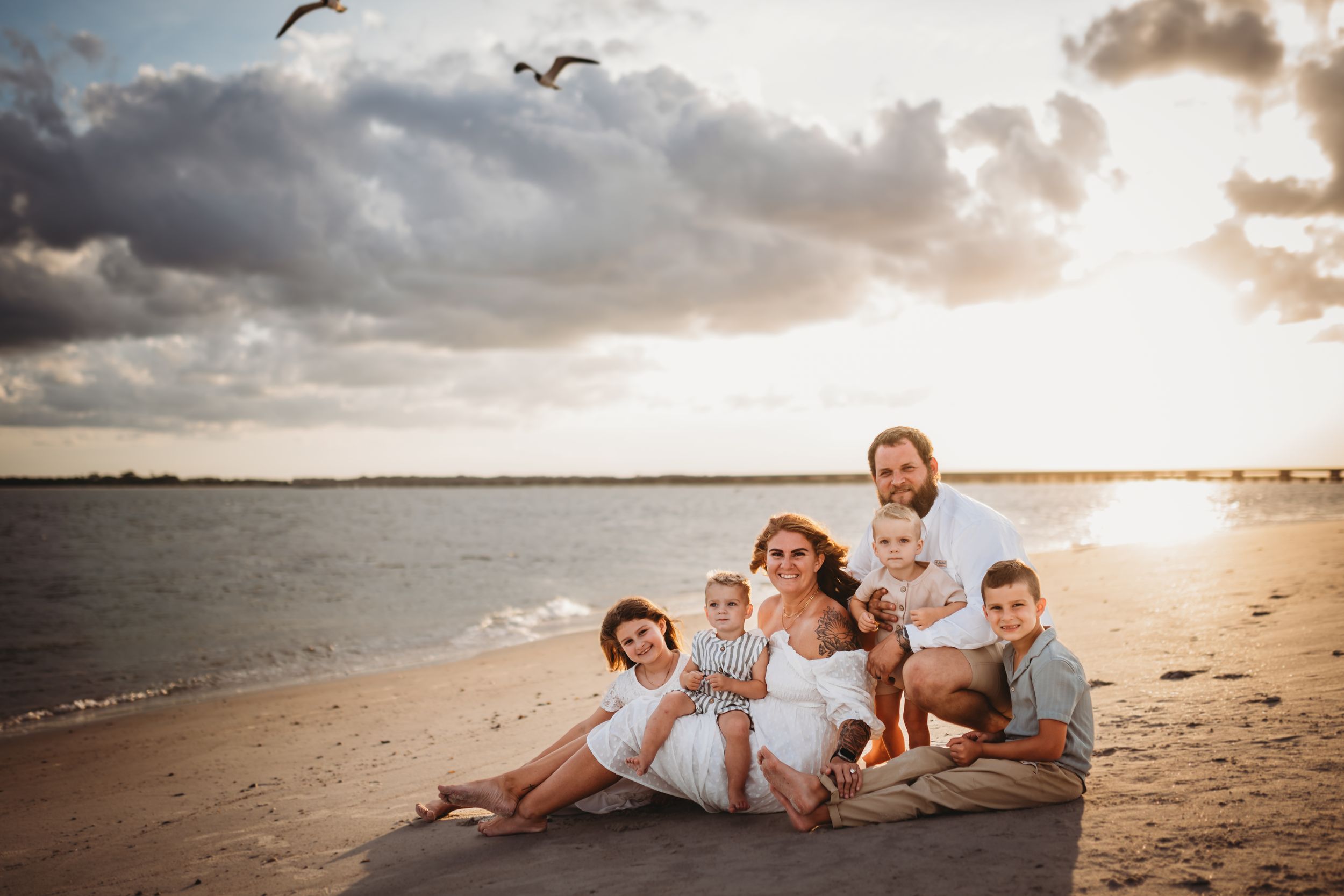 A family in white outfits sits together on a beach at sunset with seagulls flying overhead and golden sunlight.