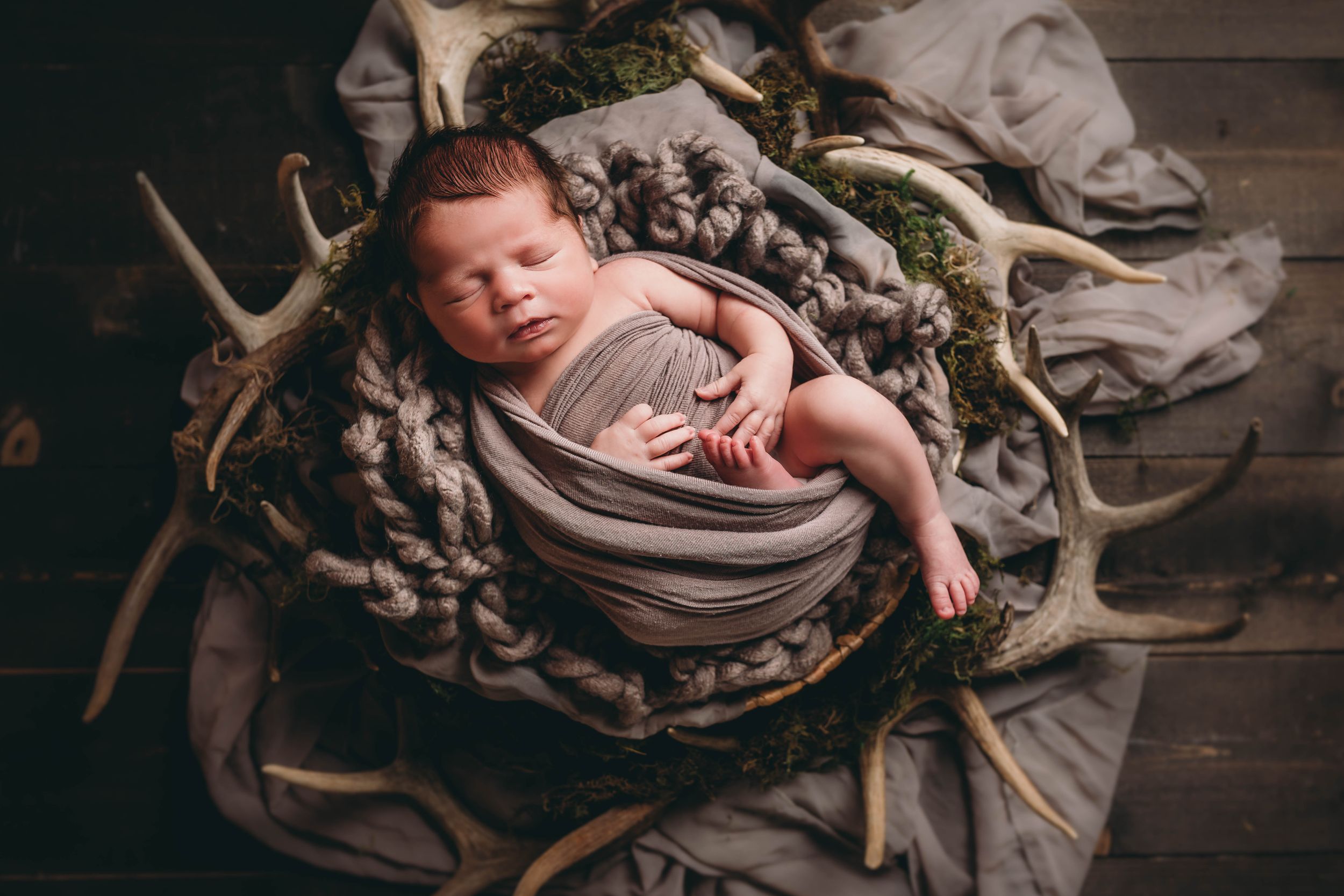 A newborn baby wrapped in soft fabric sleeps peacefully in a rustic wooden bowl surrounded by antlers and greenery.