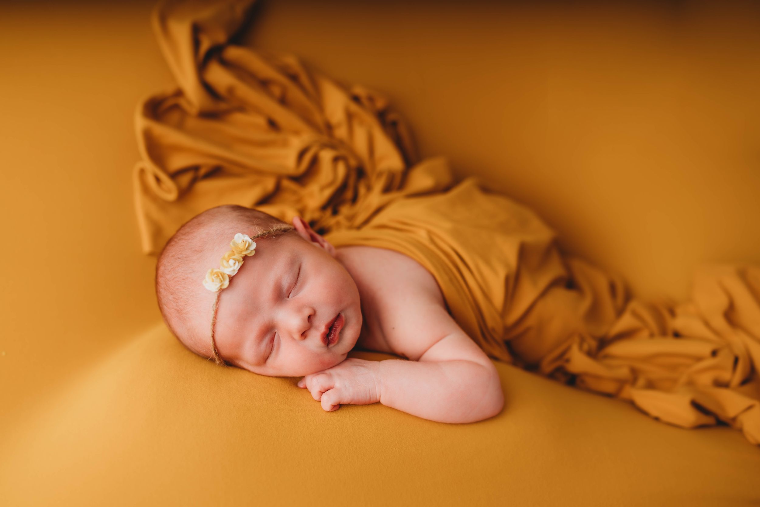 Sleeping newborn baby wrapped in soft golden fabric on warm yellow background during professional photo session.
