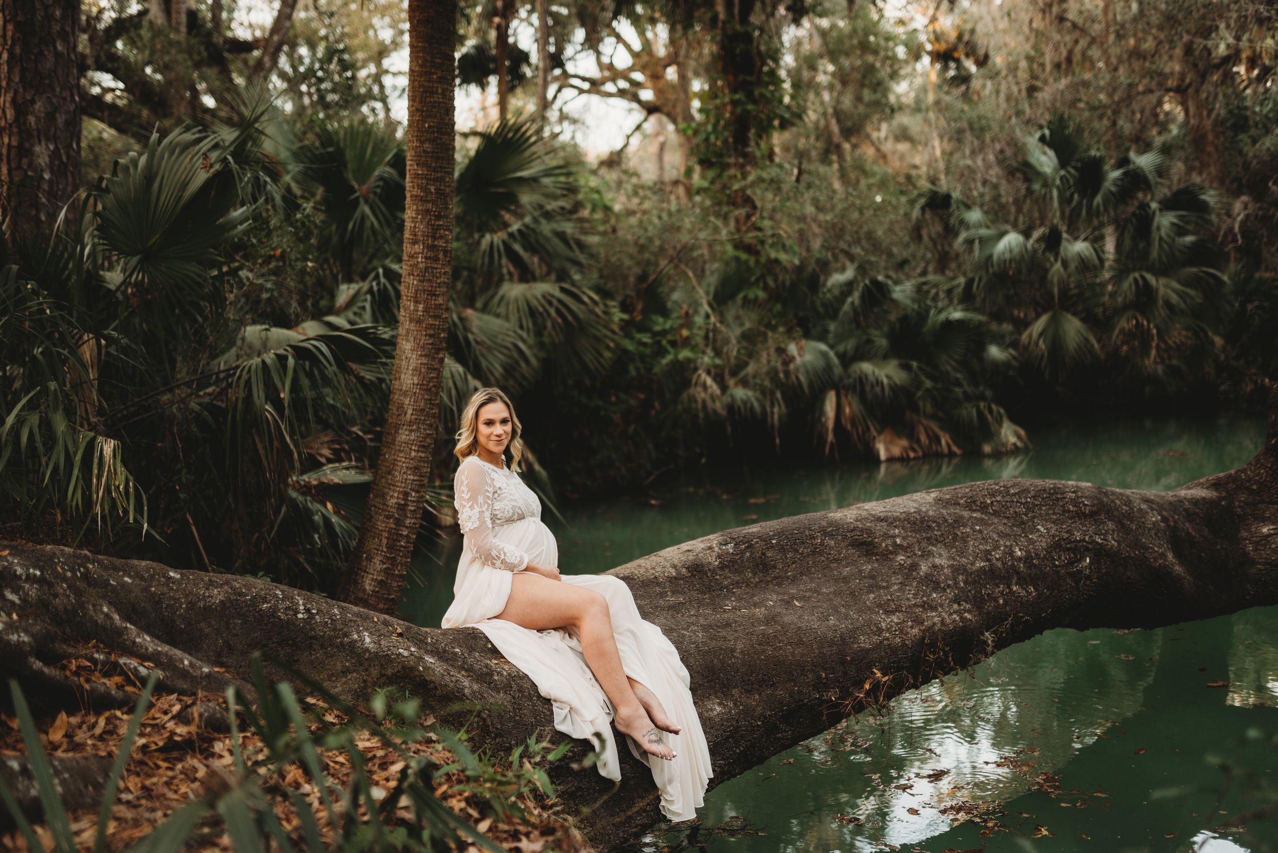Someone in a flowing white dress sits gracefully on a curved tree branch over a tranquil pond in a lush garden setting.