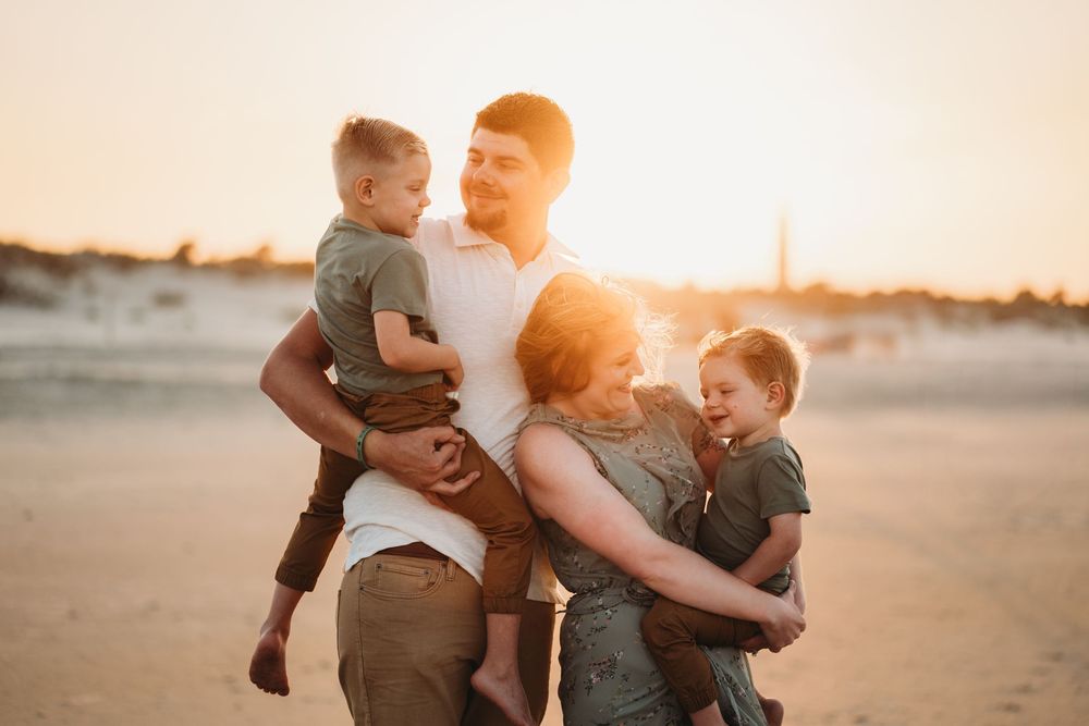 Family playing and laughing together on sandy beach at sunset with warm golden light behind them.