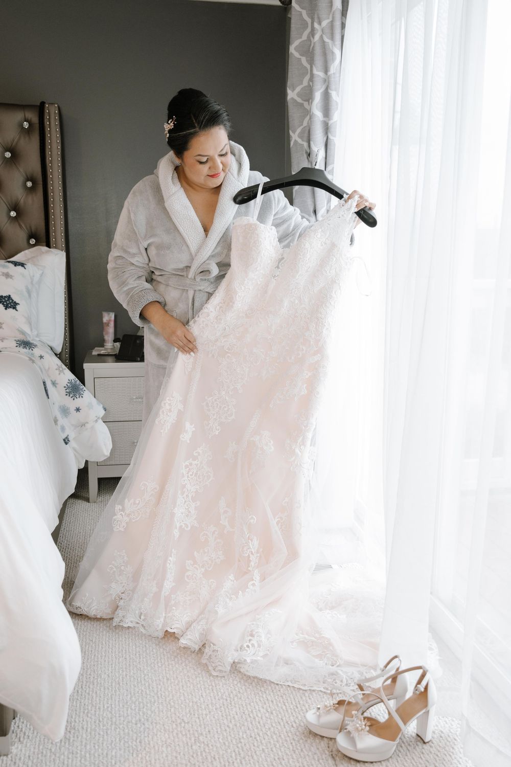 A lace wedding dress hangs by a window while someone in a robe examines the delicate details, with bridal shoes on the floor.