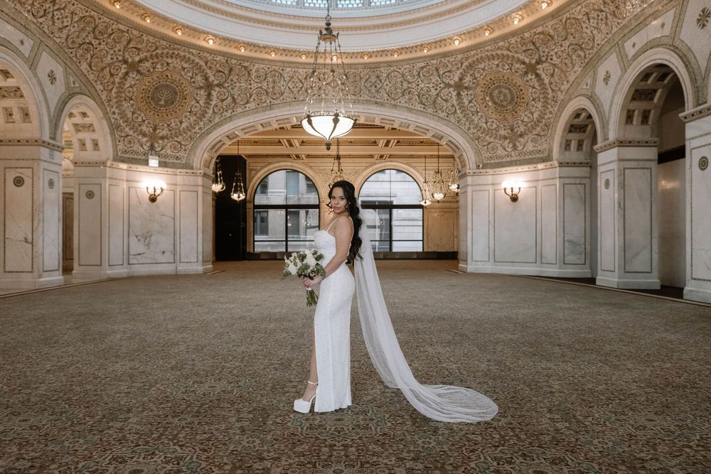 Elegant white wedding gown with flowing cape photographed in ornate historic ballroom with decorative ceiling and arches.