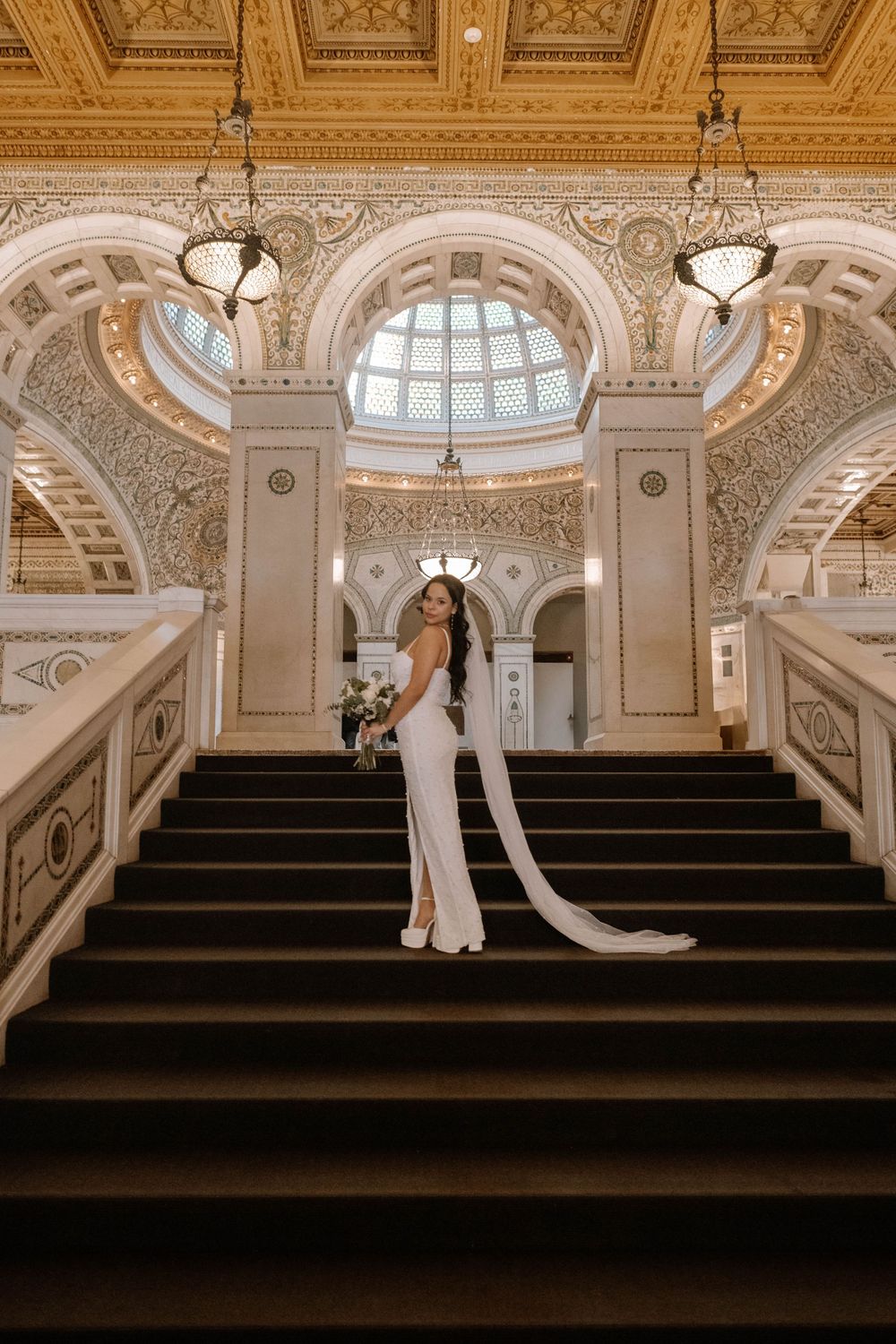 A bride in an elegant white gown with long train poses on ornate marble staircase under domed ceiling with chandeliers.