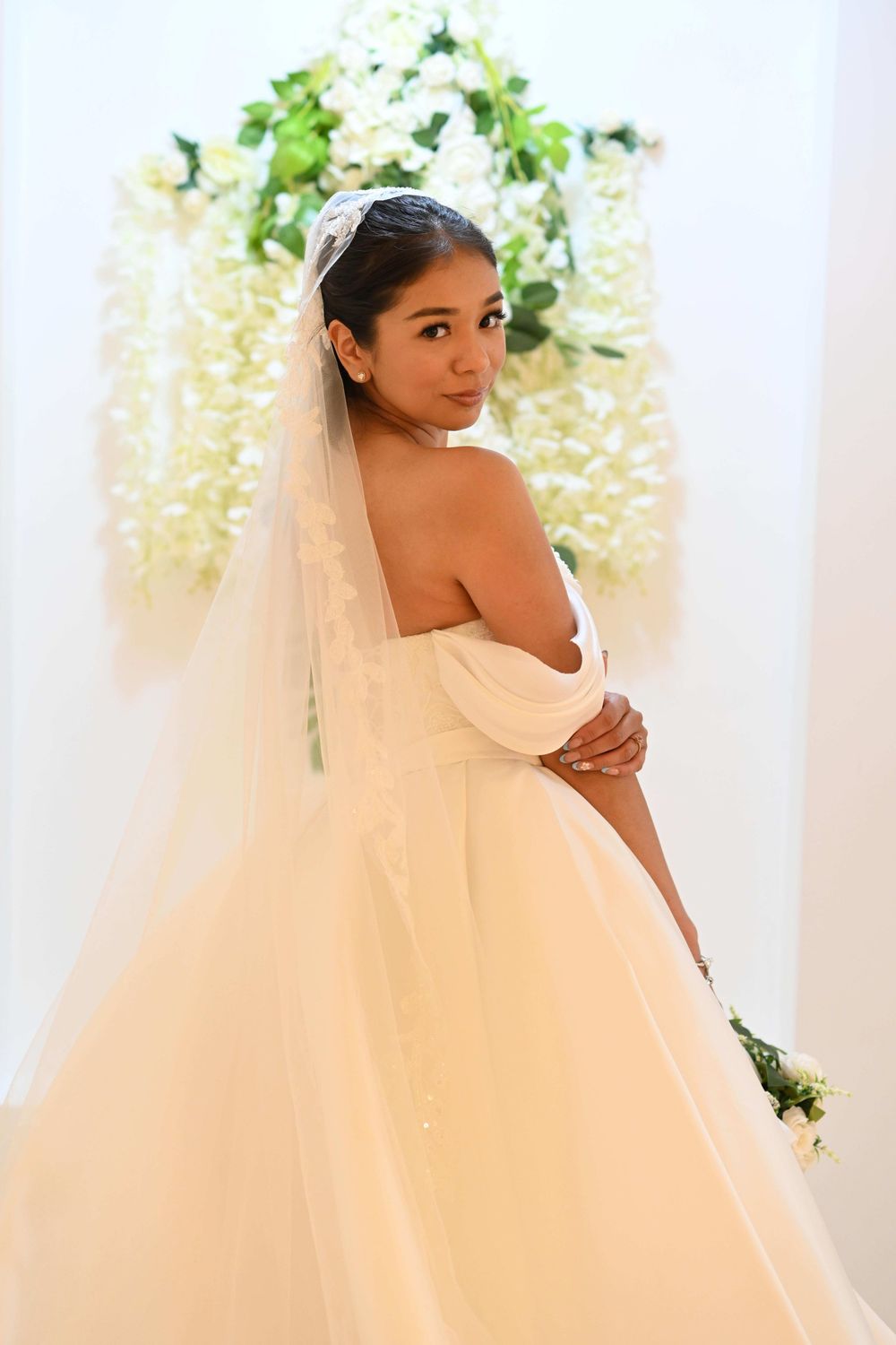 A bride in a stunning off-shoulder white ball gown and veil poses elegantly against a backdrop of floral decorations.
