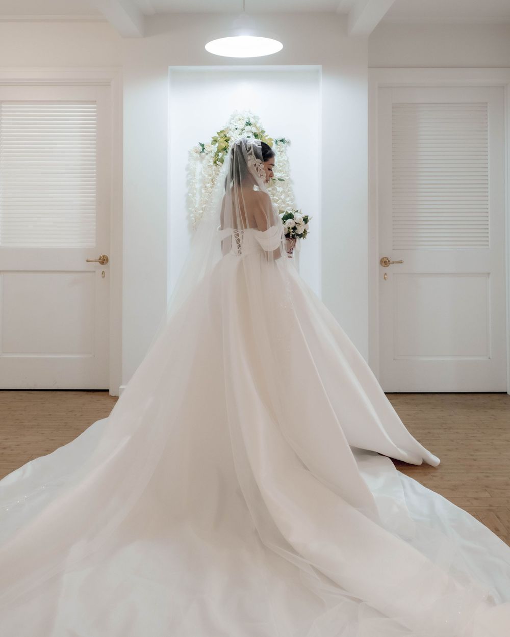 Elegant bride in flowing white wedding gown with long train and veil stands between two doors in bright hallway.
