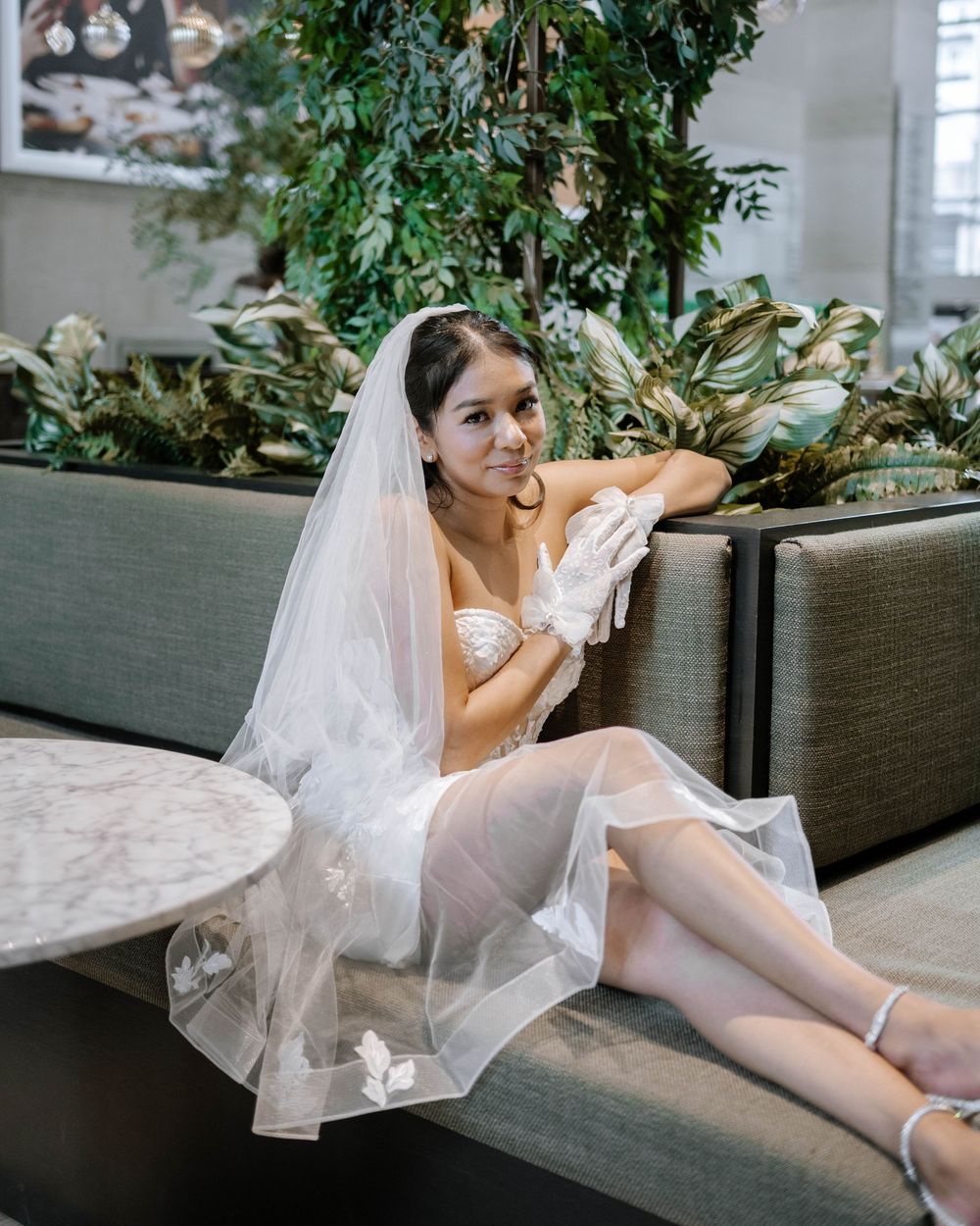 A dreamy bridal portrait on a modern couch surrounded by lush greenery and soft lighting in an indoor setting.