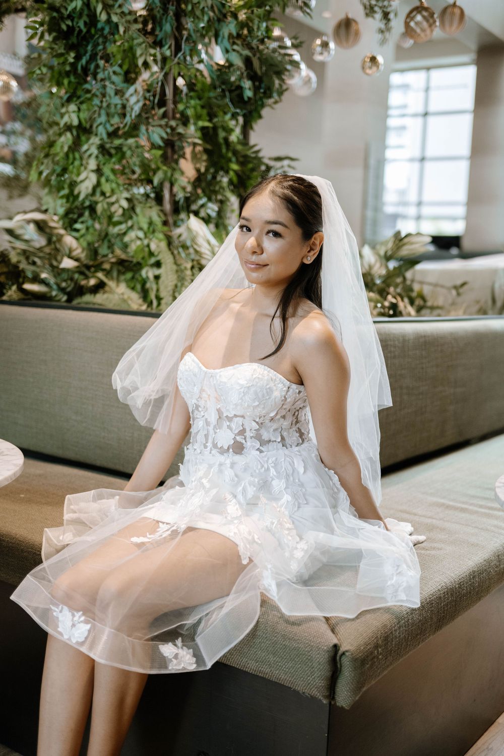 Elegant white lace wedding gown with sheer tulle detail sits gracefully on a grey couch in a bright sunlit room with plants.
