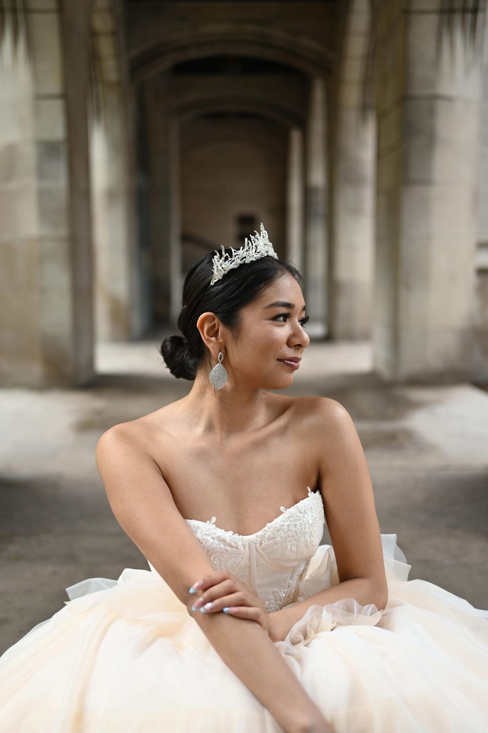 Elegant bride in a strapless white ball gown and tiara sits gracefully in an arched stone corridor.