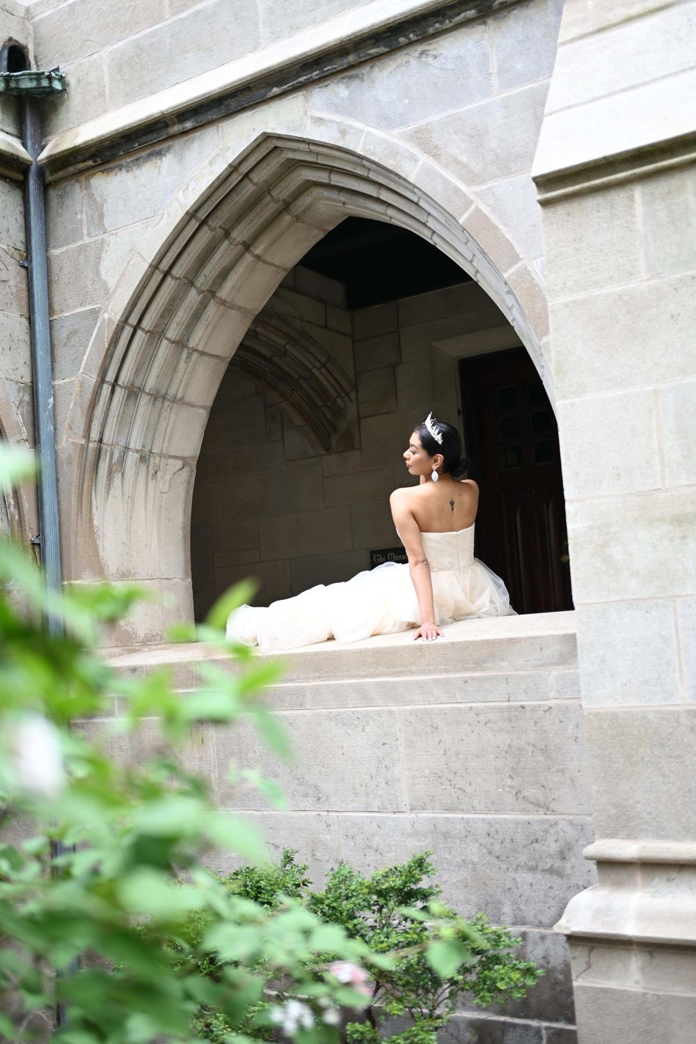A figure in a white gown sits at an arched stone window ledge of a historic Gothic-style building framed by greenery.