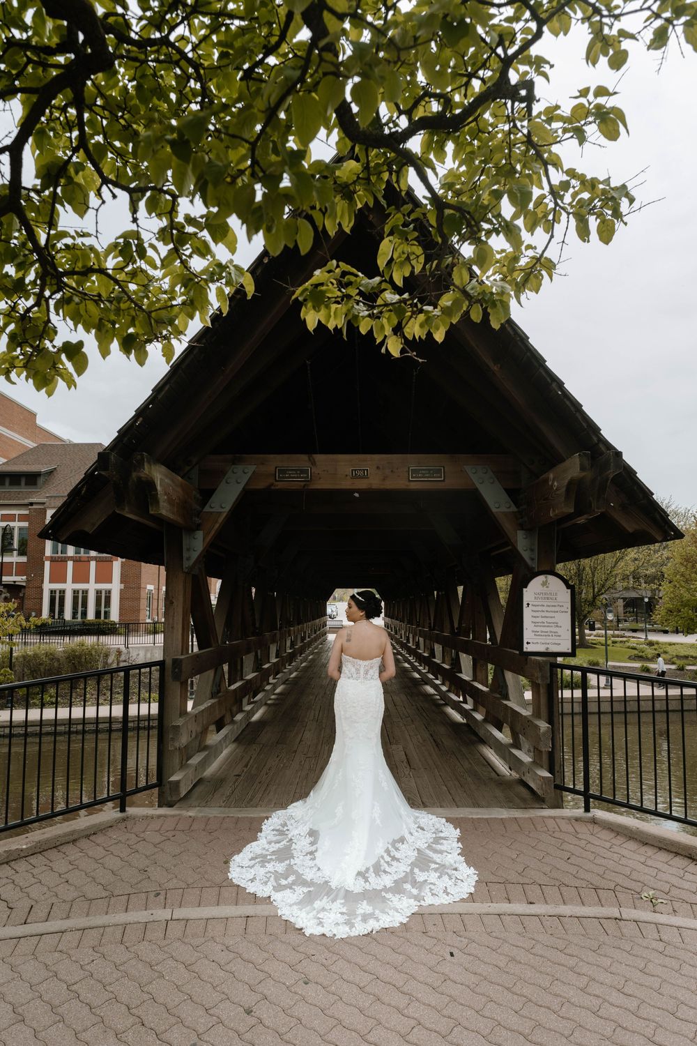 A white lace wedding dress with a dramatic train photographed on a covered wooden bridge surrounded by green foliage.