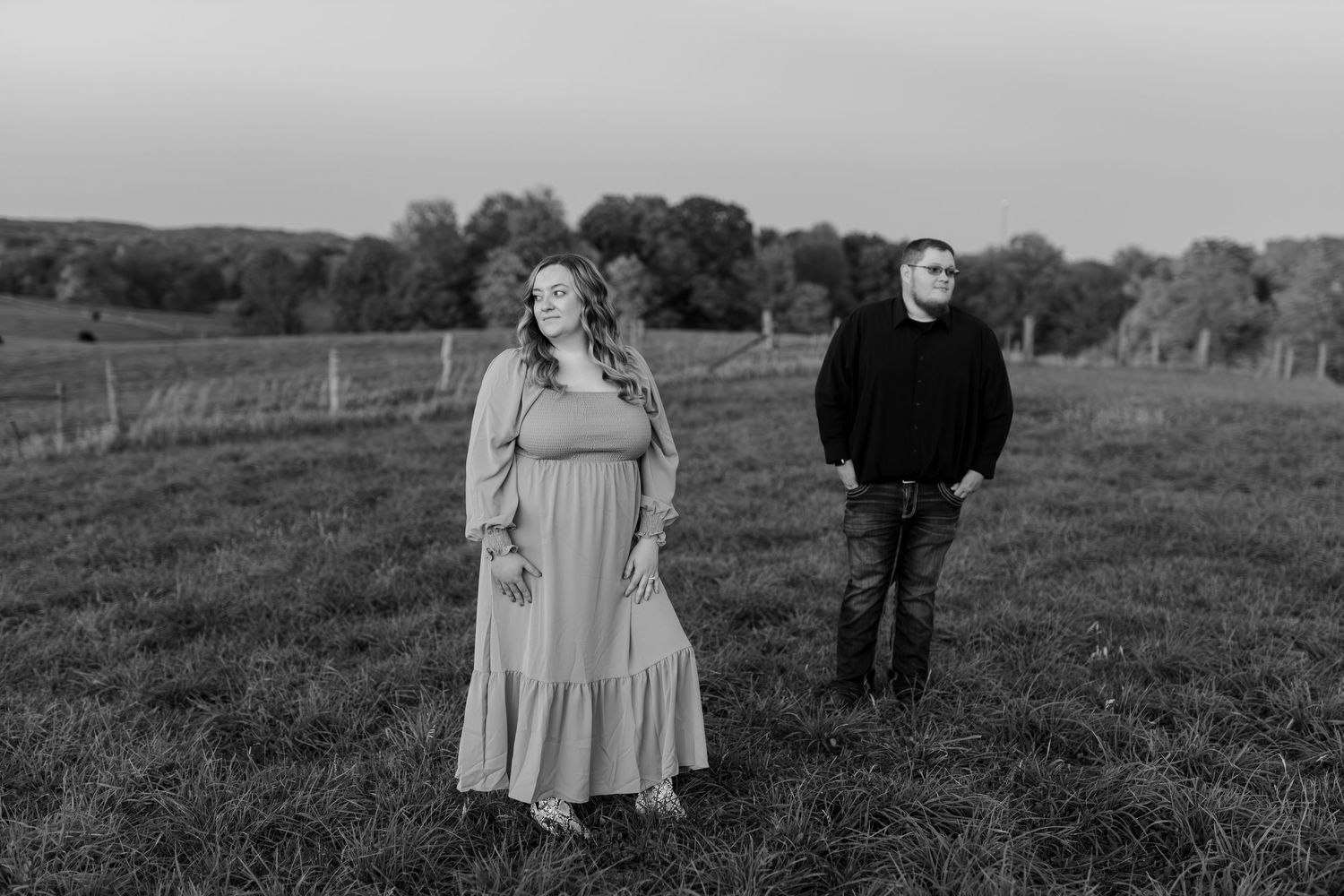 Black and white engagement photo of a couple standing apart in a lush green field at dusk.