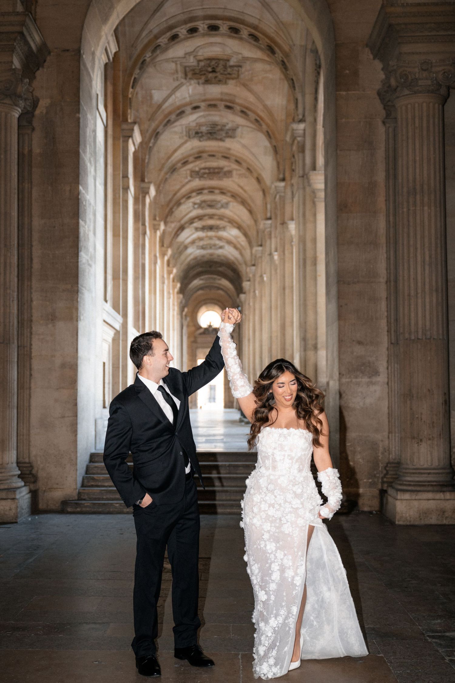 A couple dancing together under elegant stone archways with palm trees visible in the background.