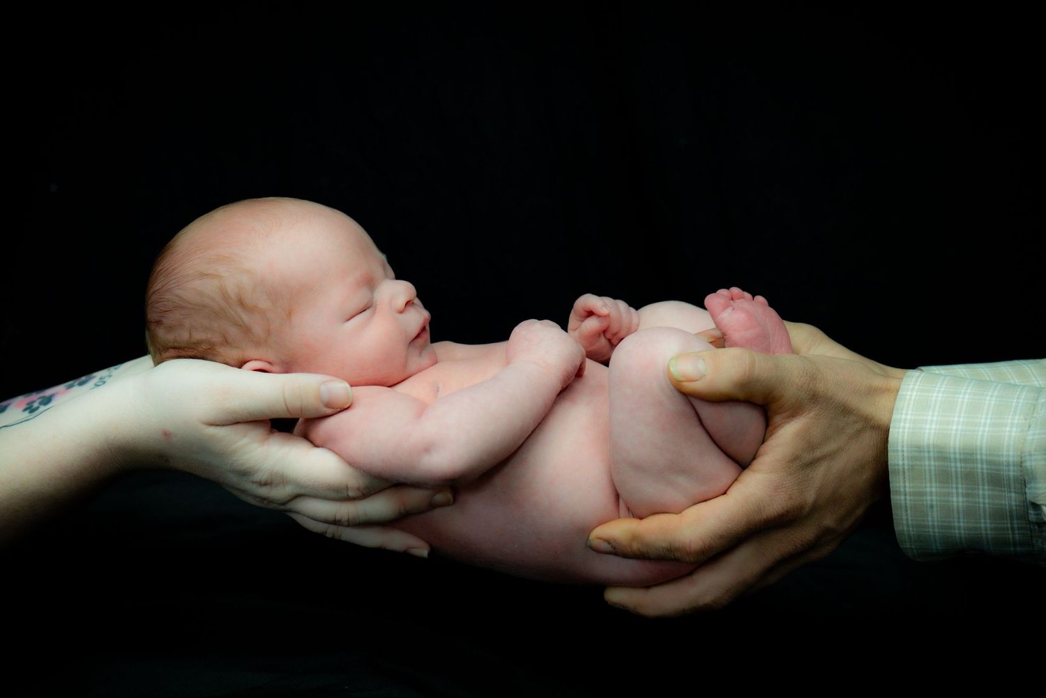 A newborn baby cradled between adult hands against a black background.