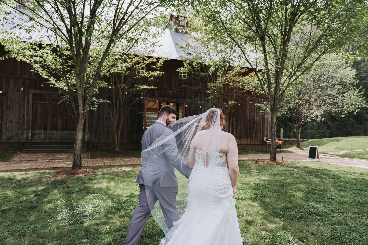 A couple in wedding attire walk together on a sunny lawn with trees and a rustic wooden building in the background.