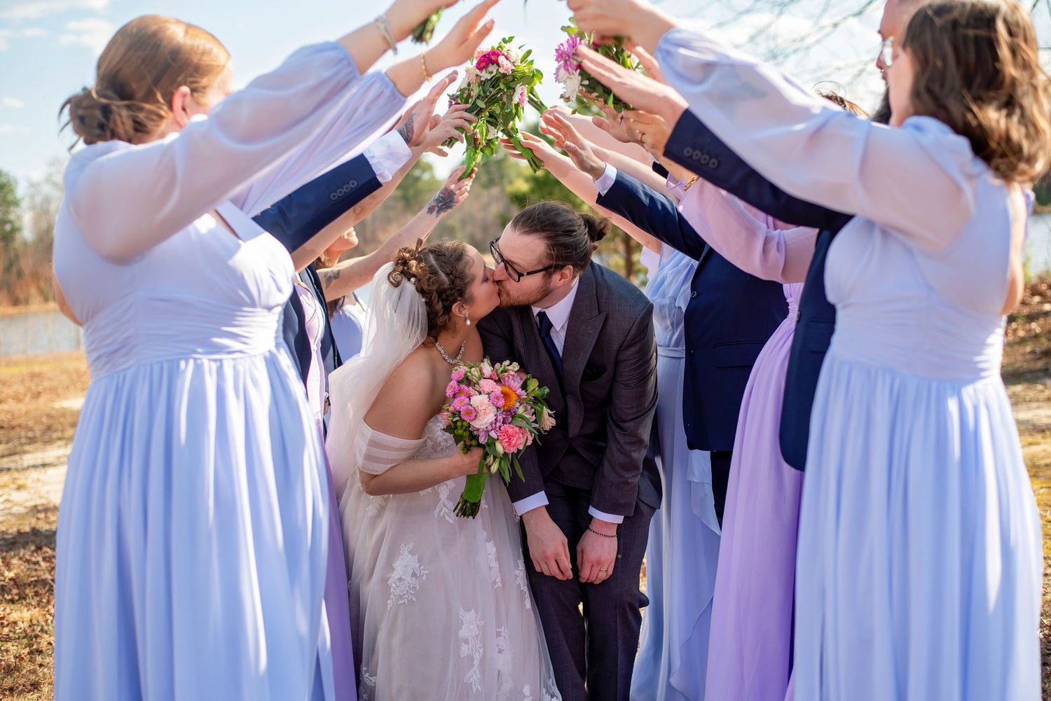 Wedding party in light purple dresses form archway over couple during outdoor ceremony with mountain backdrop.