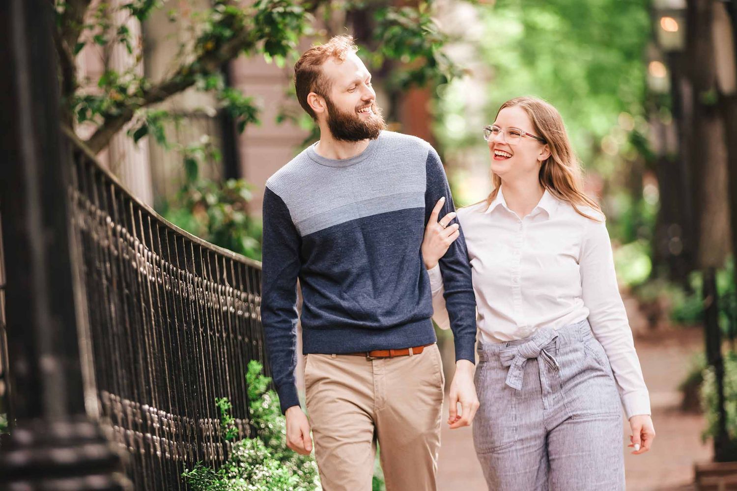 A joyful couple laughs together while strolling past elegant wrought iron fences and blooming plants on a Beacon Hill sidewalk