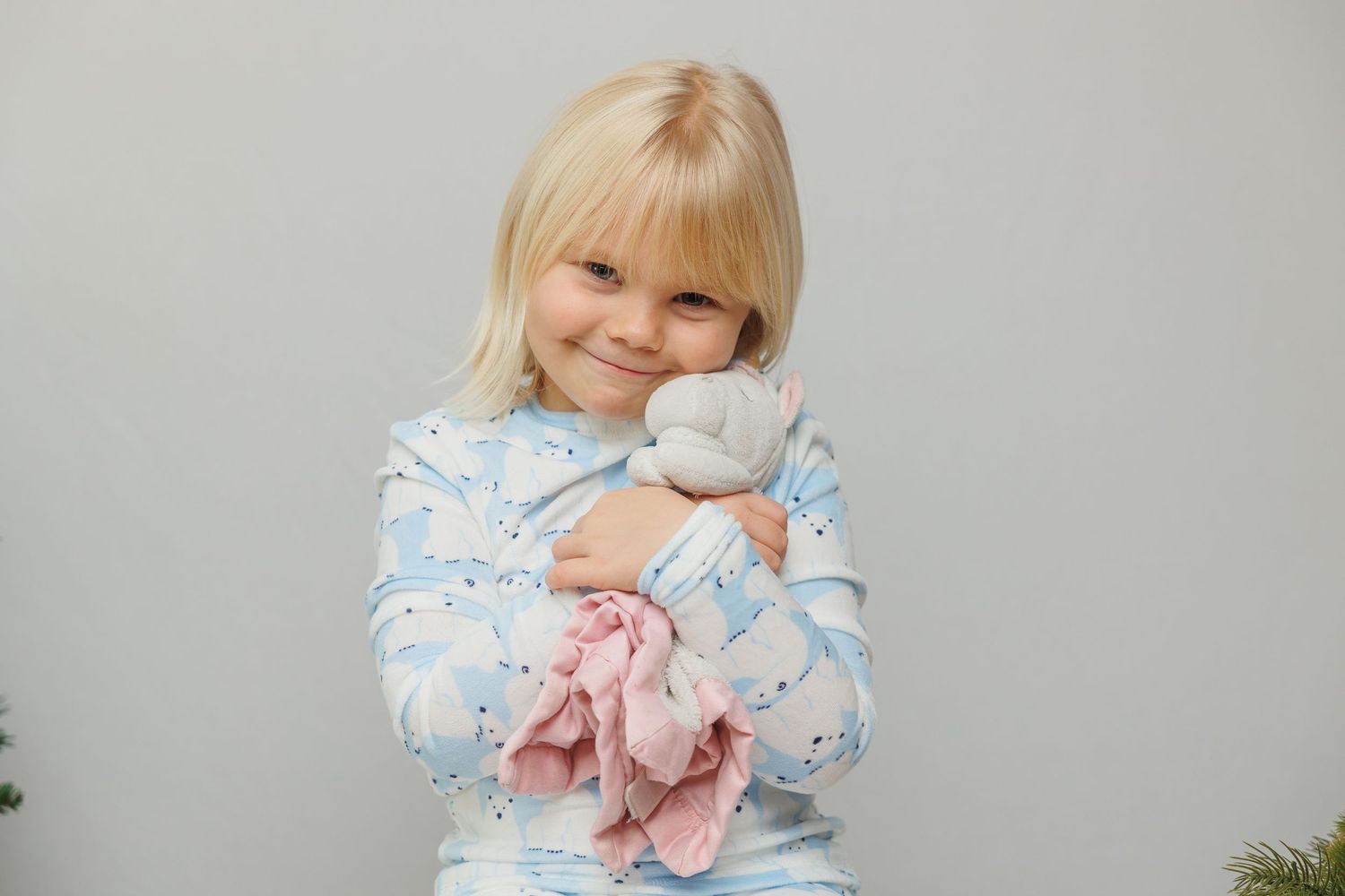 Young child in blue floral sleepwear holding a soft pink stuffed toy against a gray background.