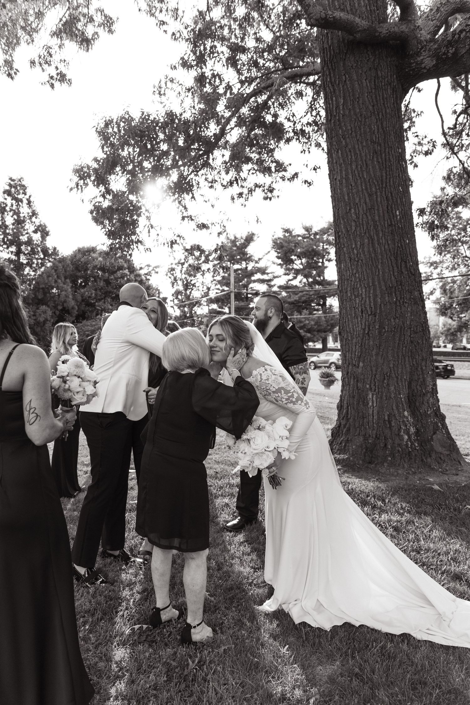 Wedding party gathers under a large tree outdoors as guests embrace the person in a long white gown with flowing veil.