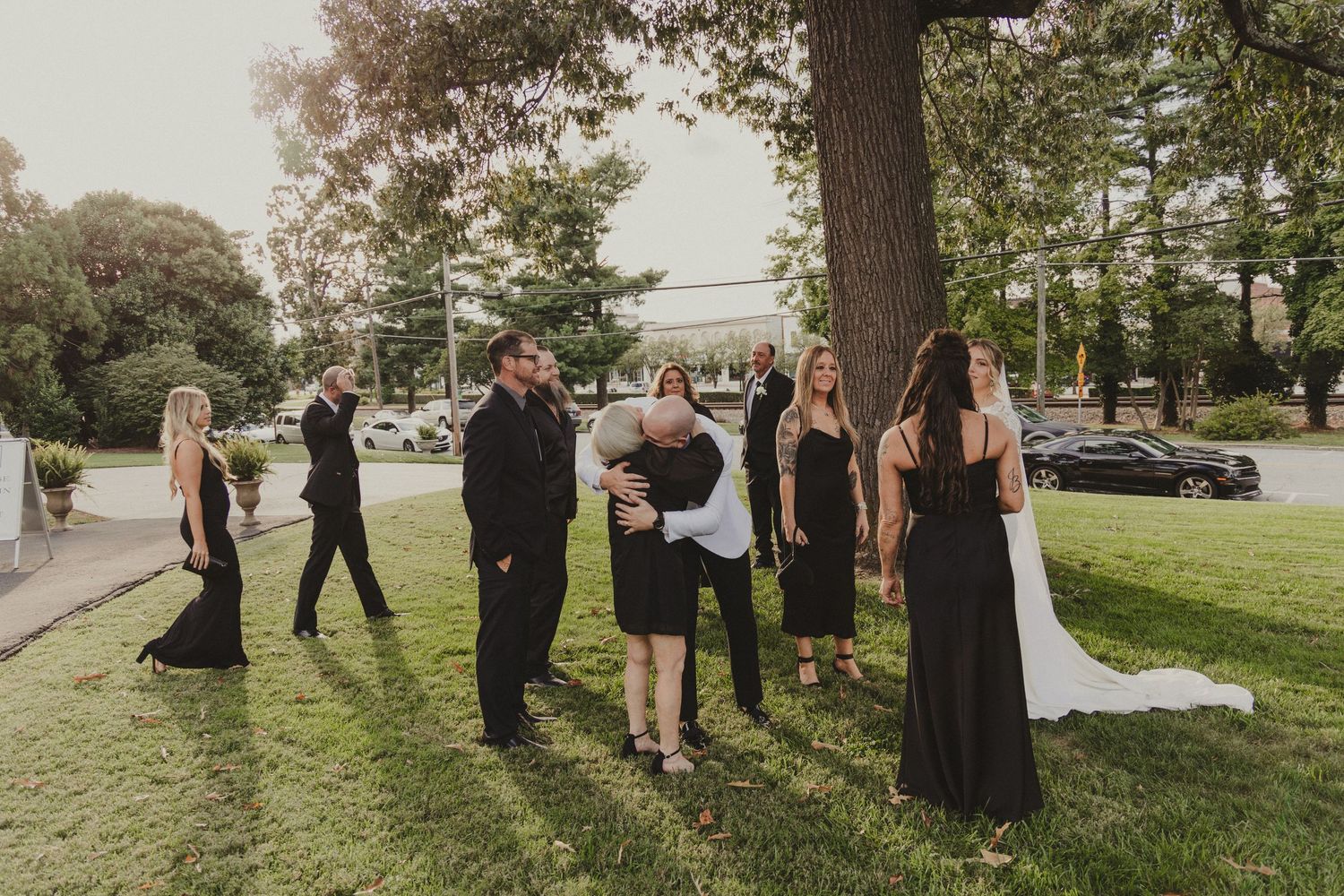 A group of formally dressed people gather outdoors on a grassy lawn surrounded by trees at a special event.