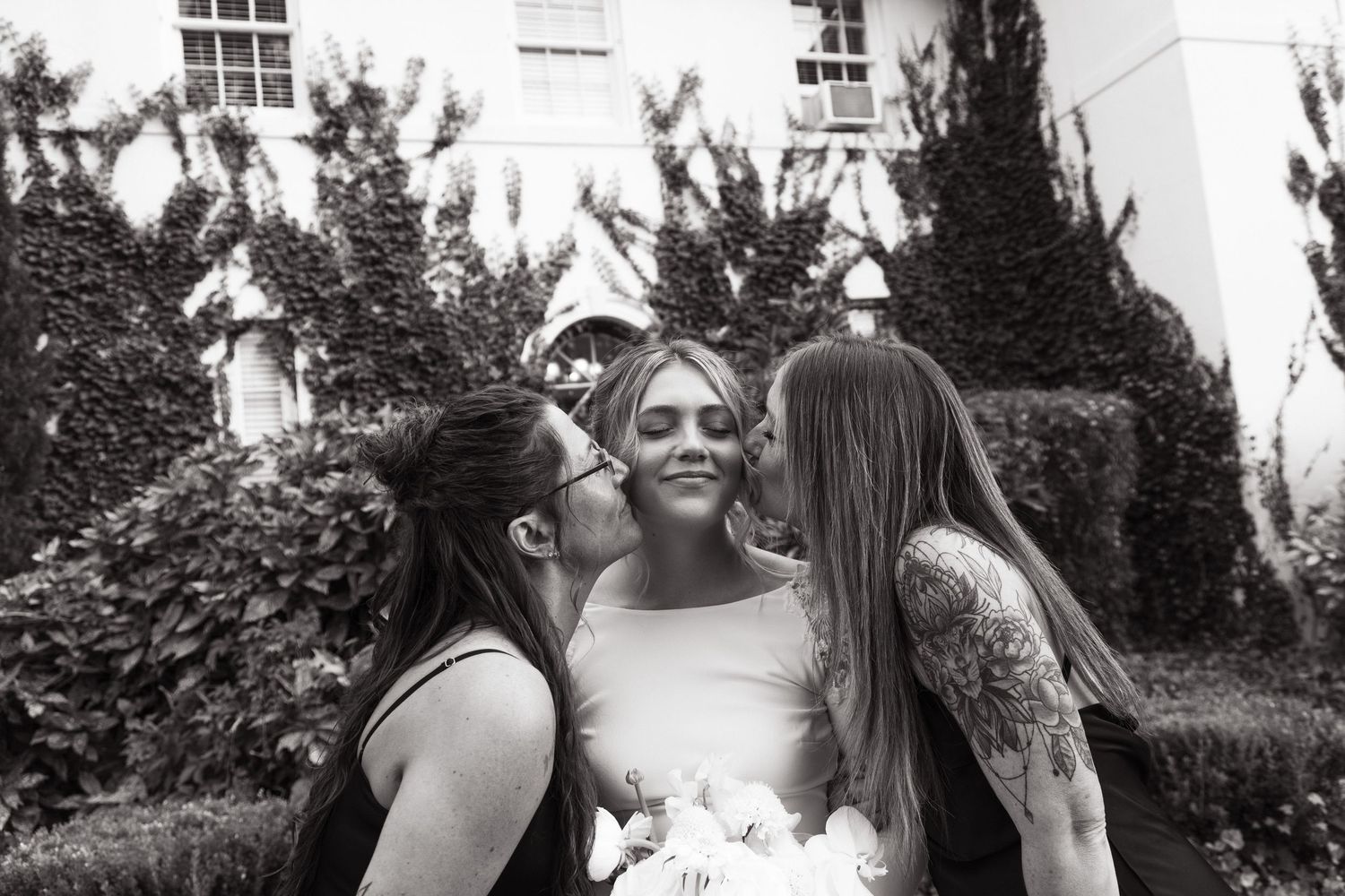 A black and white photo shows three people sharing an intimate moment in a garden setting with bushes and white walls.