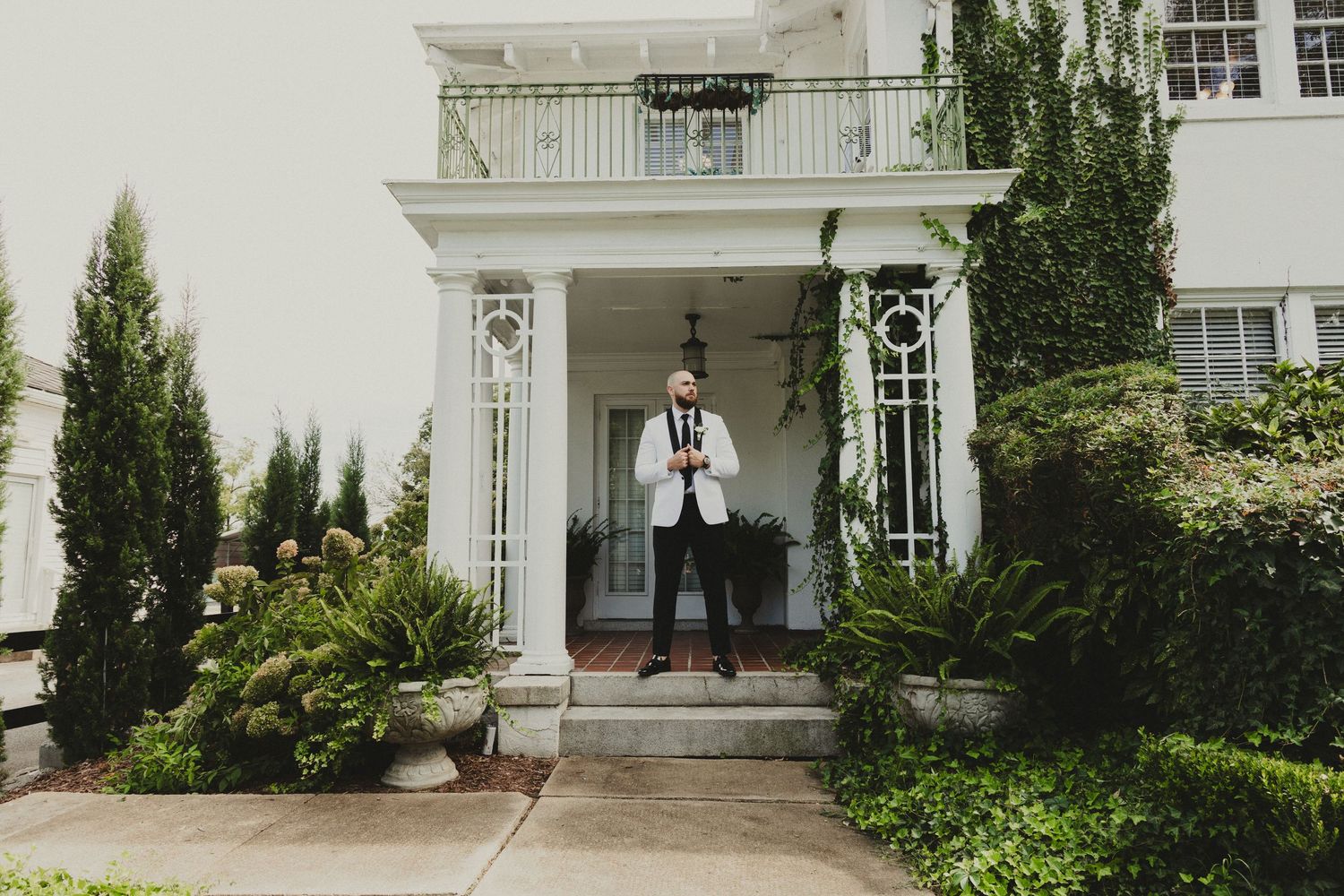 A person in formal attire stands on a white columned porch entrance surrounded by lush greenery and landscaping.