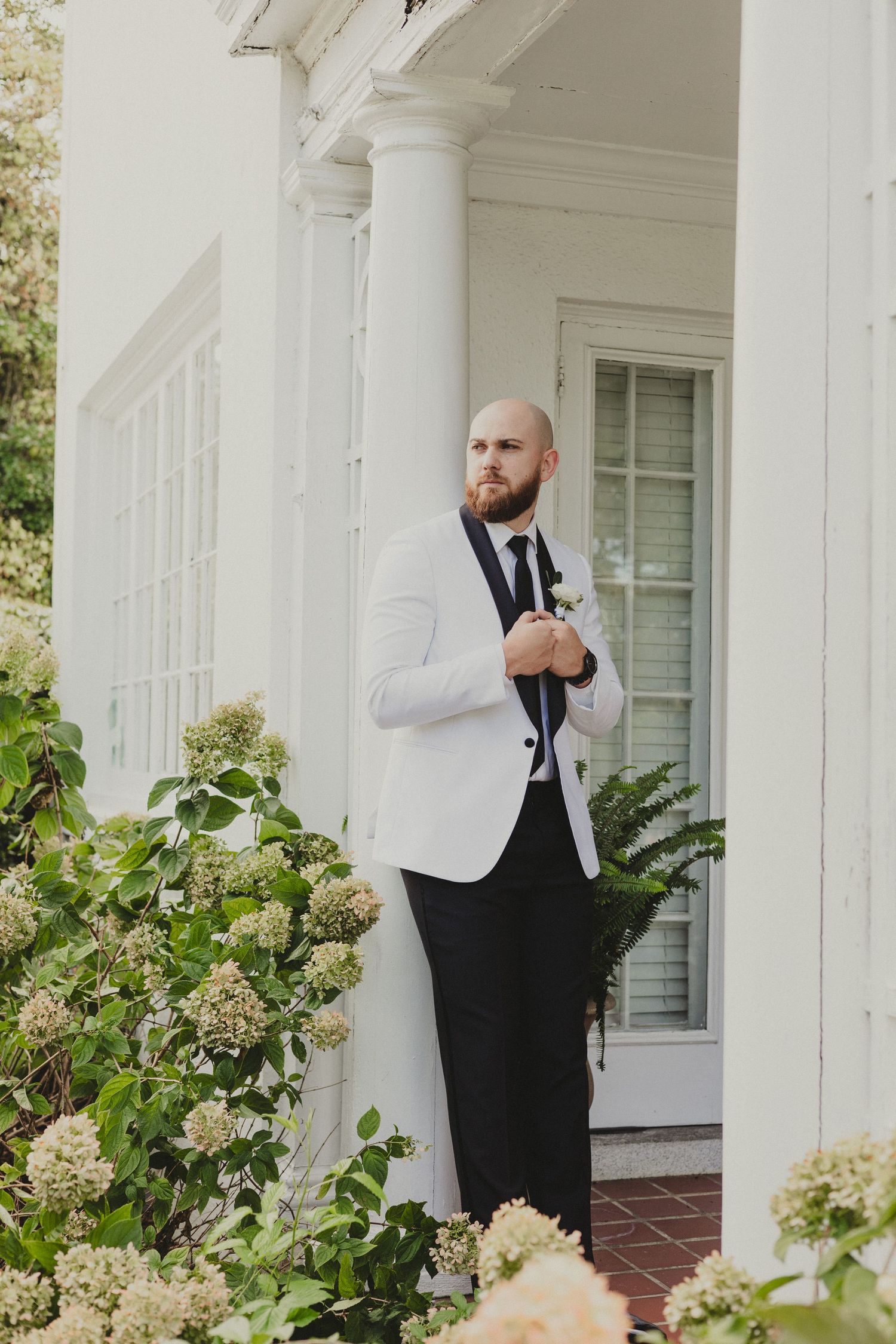 A groom in a white tuxedo jacket and black pants stands on an elegant white porch surrounded by flowering hydrangea bushes.