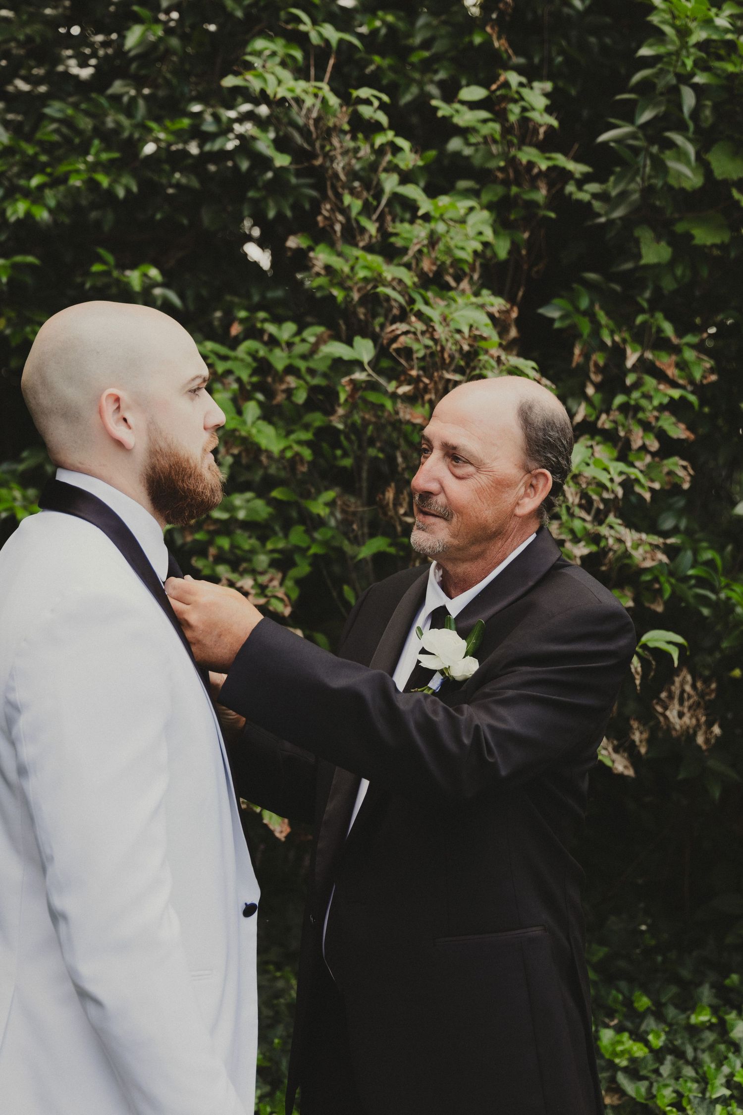 A tender moment between two well-dressed gentlemen adjusting formal attire against a leafy green backdrop at a wedding ceremony.