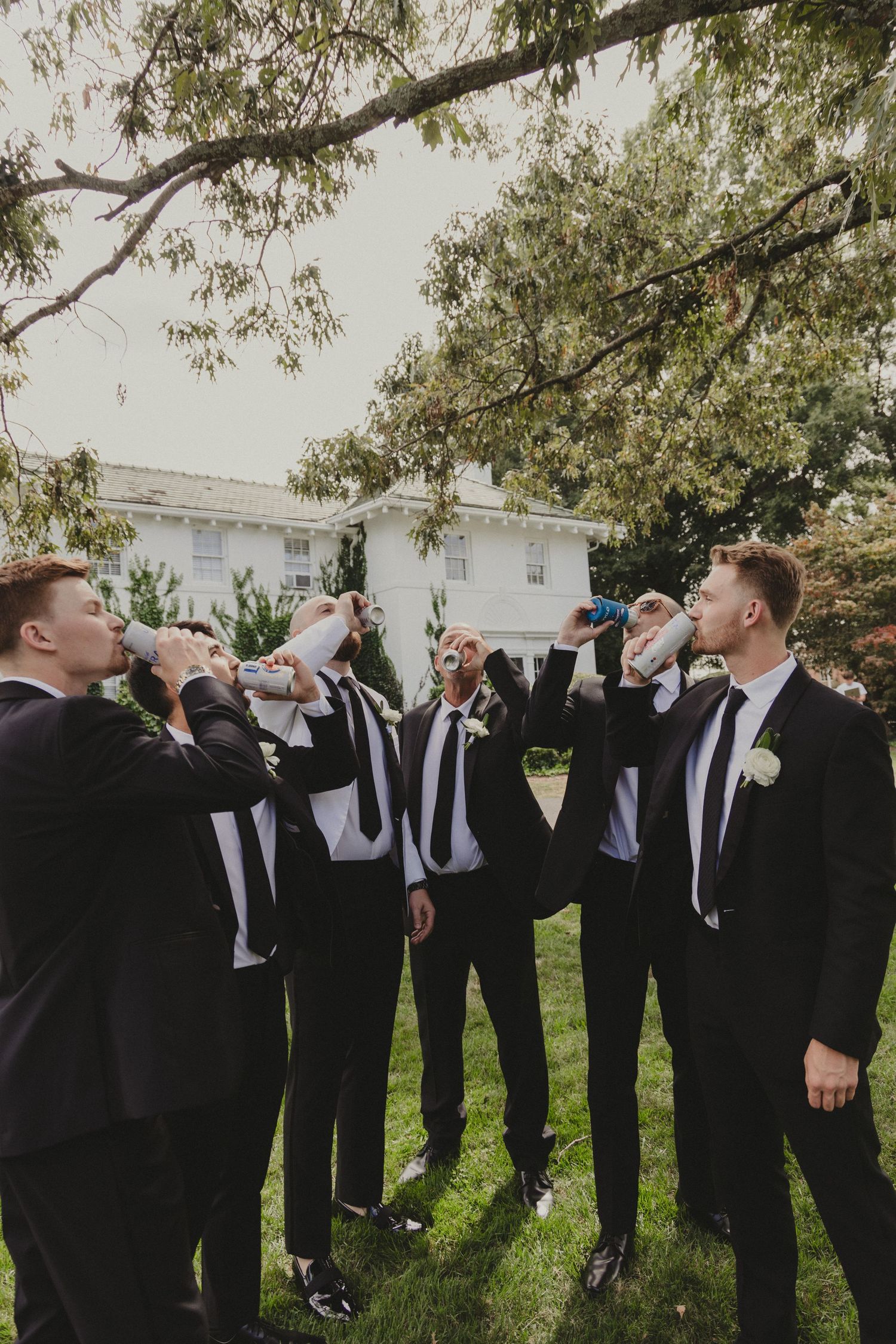 Groomsmen in black suits drink together while standing on grass lawn near white building during wedding celebration.