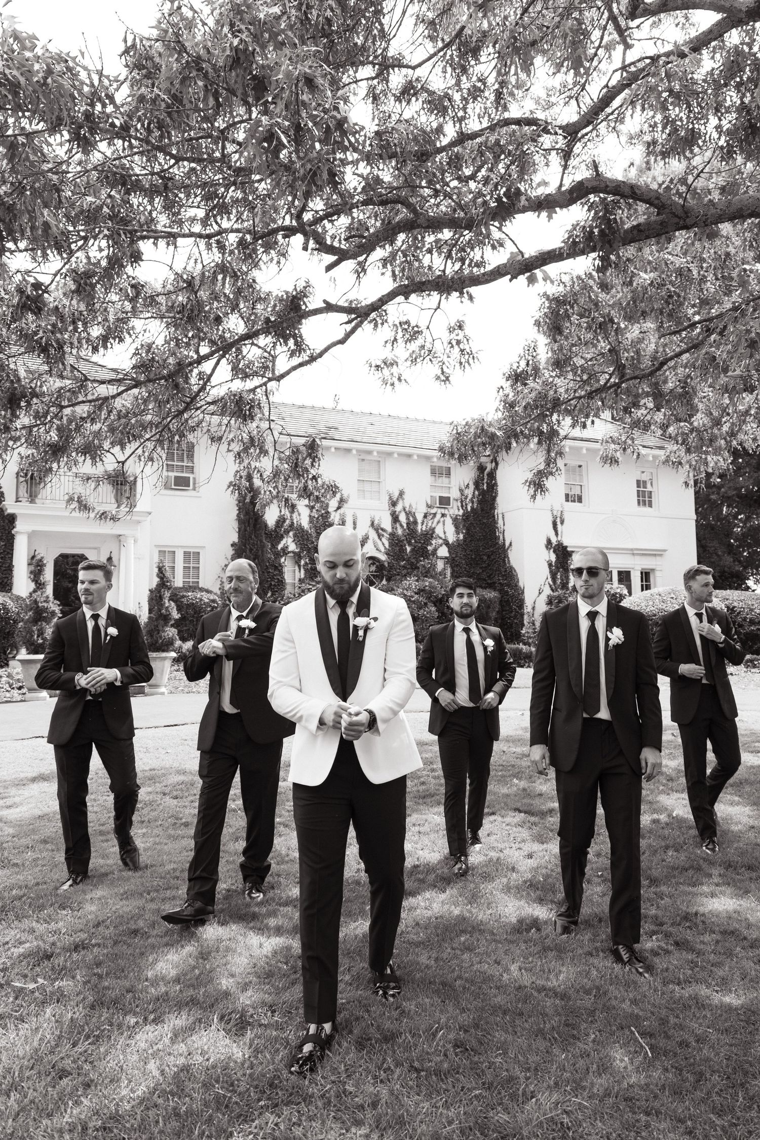Black and white photo showing a wedding party with groomsmen in dark suits behind one person in a white jacket outdoors.