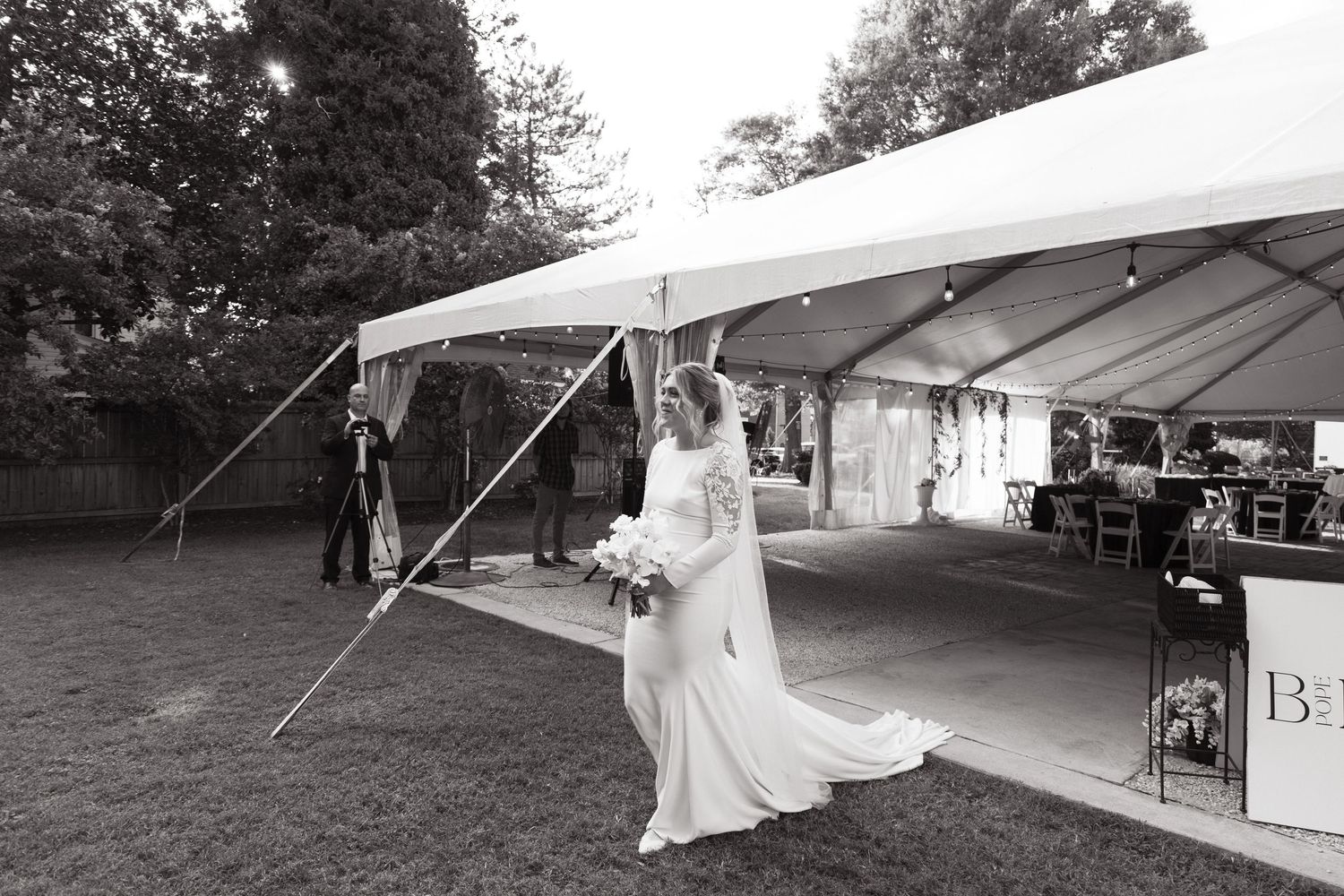 A black and white outdoor wedding photo under a white tent with a bride in a gown and guests on a lawn.