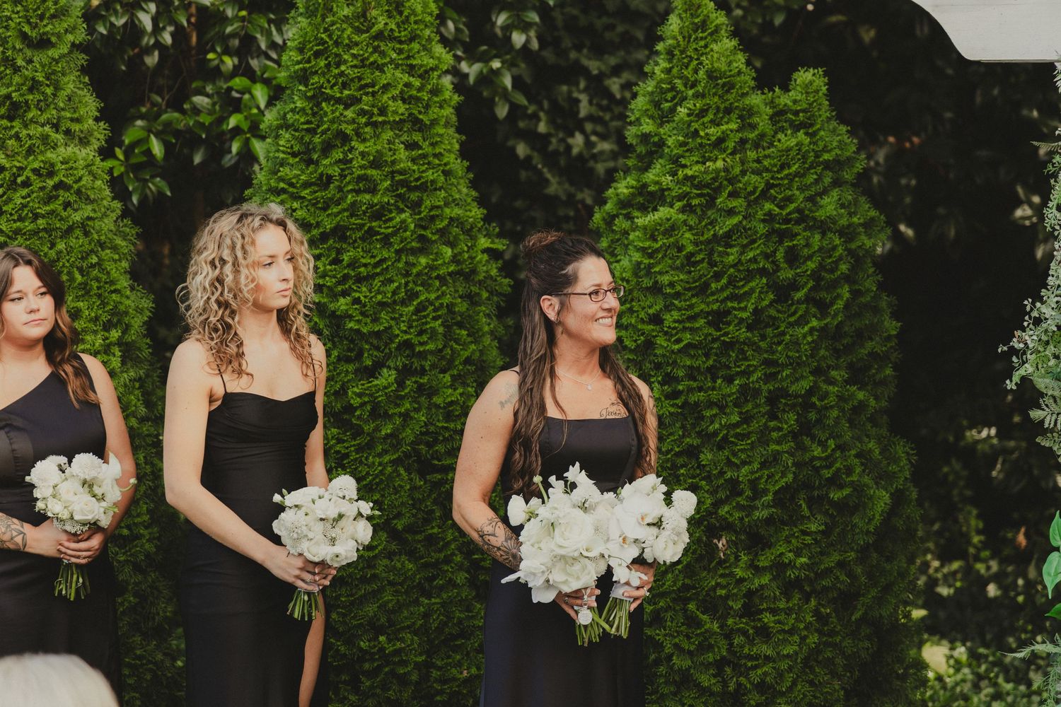 Bridesmaids in elegant black dresses hold white floral bouquets while standing in front of manicured evergreen bushes.